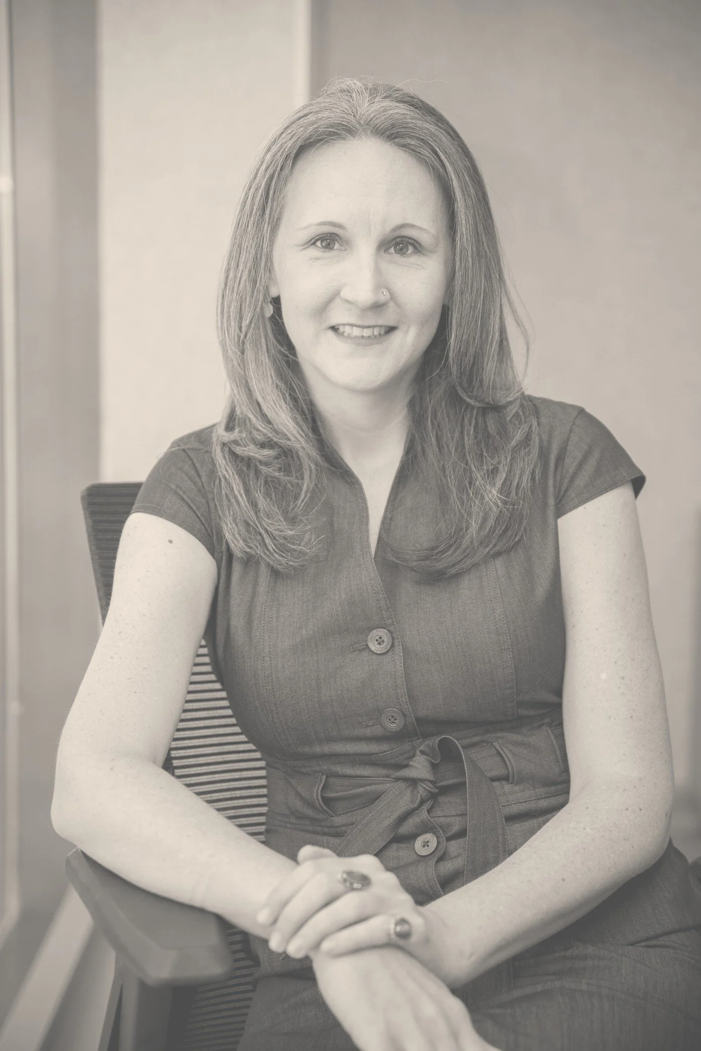A woman with dark brown hair wearing a pink floral blouse, sitting at a desk with a book, a mug, and a black office chair, smiling.