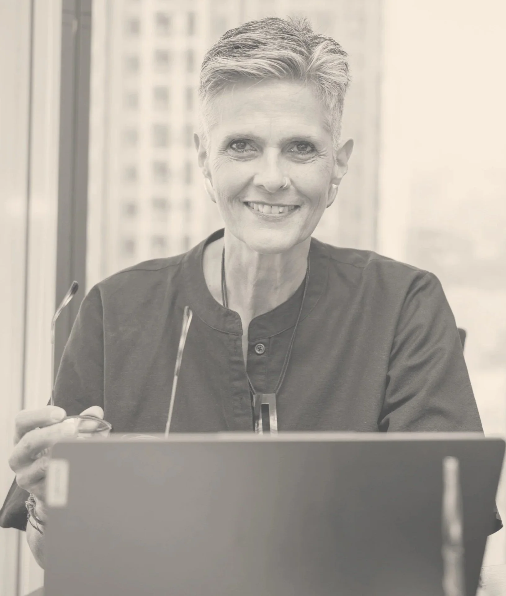 A mature woman with gray hair and glasses, wearing a patterned dress, pearl necklace, and pearl earrings, sits at a desk with her hands clasped, looking confidently at the camera.