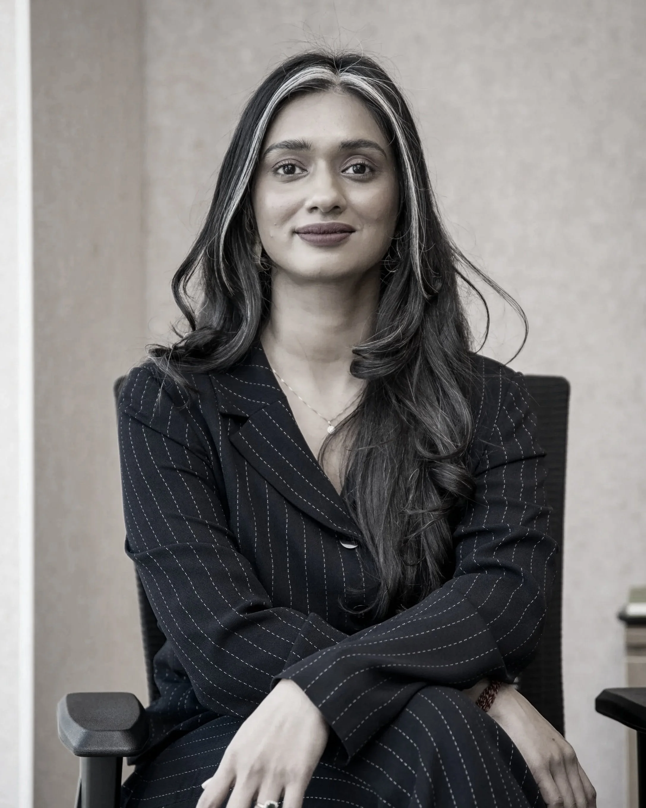 A young woman with long dark hair, wearing a yellow top, sitting at a desk, smiling, with a hand holding a pen near an open notebook in front of her.