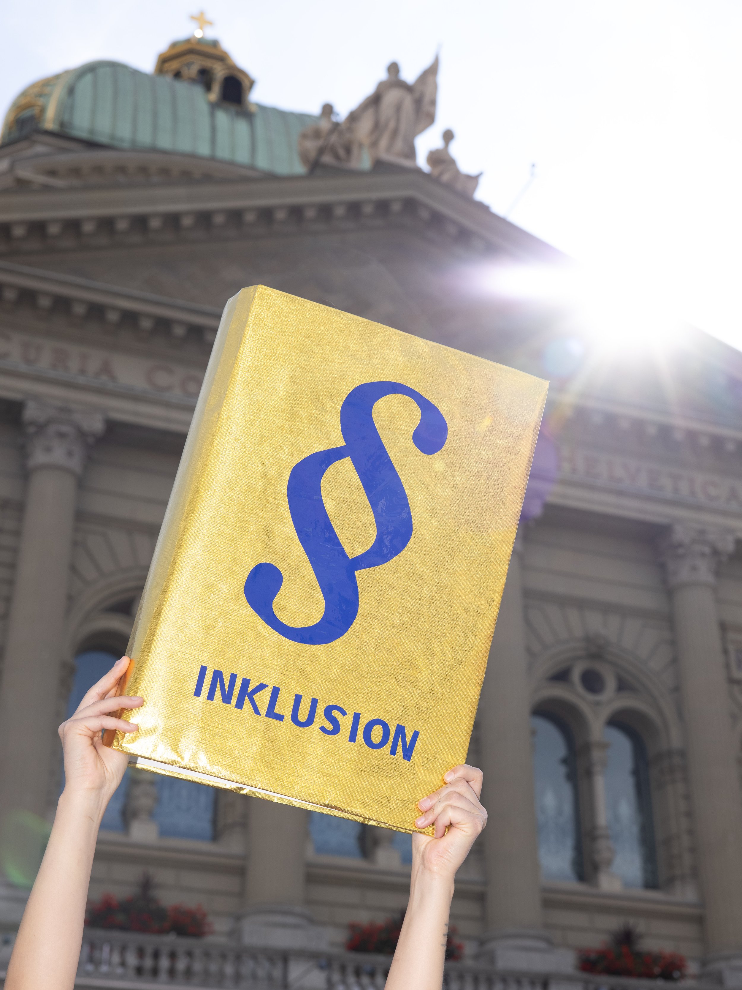 Symbolisch wird ein goldenes Buch mit einem Paragraphen-Zeichen und dem Schriftzug "Inklusion" vor dem Bundeshaus in die Höhe gehalten.