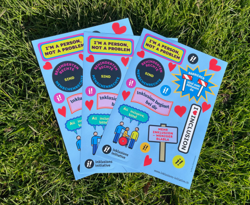 Ein Stapel von Stickerbögen mit den bunten Inklusionsstickern liegt auf einem Rasen. Die Sticker haben verschiedene Formen und Slogans, wie z.B. "Inklusion beginnt bei dir".