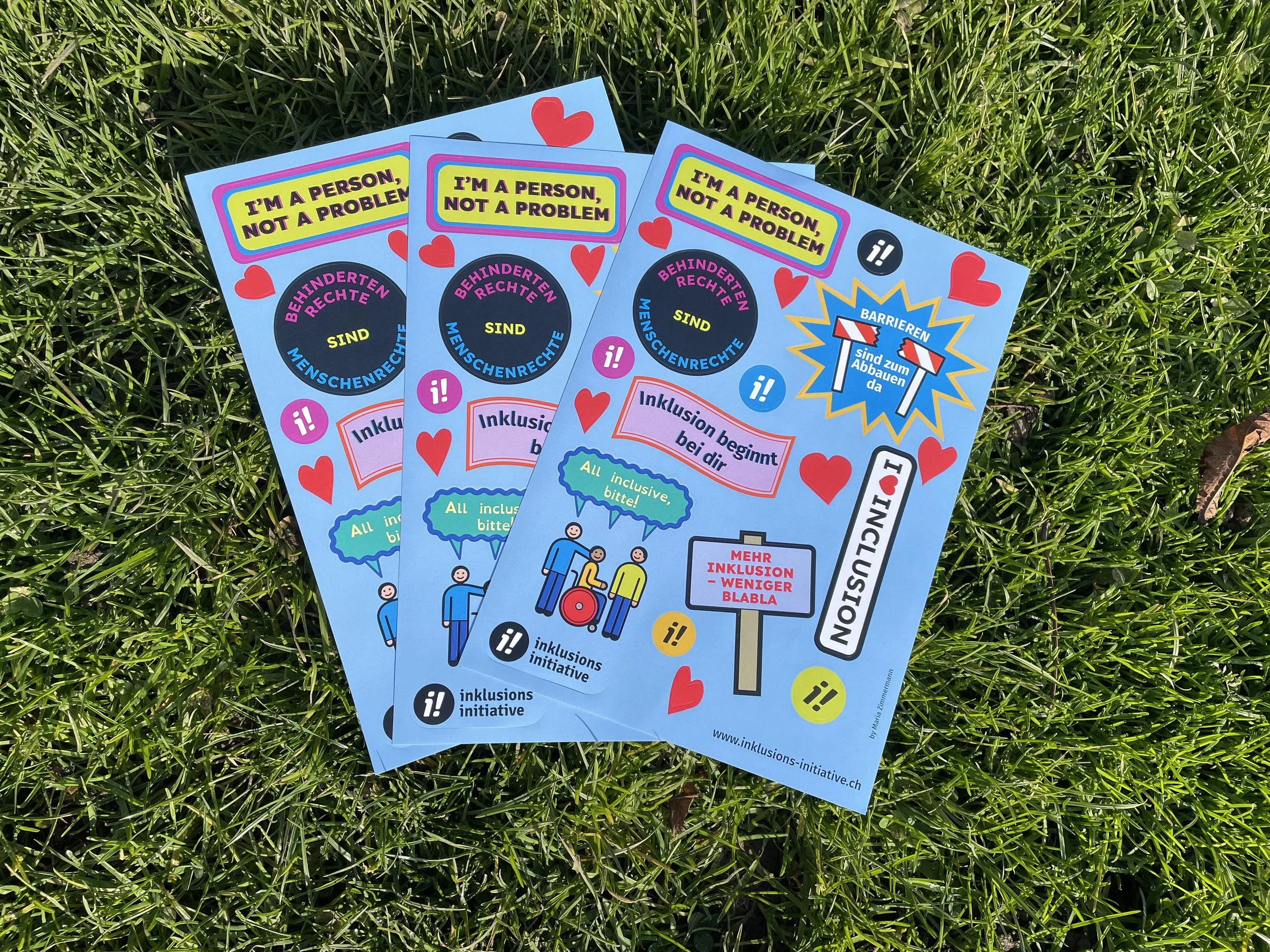 Ein Stapel von Stickerbögen mit den bunten Inklusionsstickern liegt auf einem Rasen. Die Sticker haben verschiedene Formen und Slogans, wie z.B. "Inklusion beginnt bei dir".