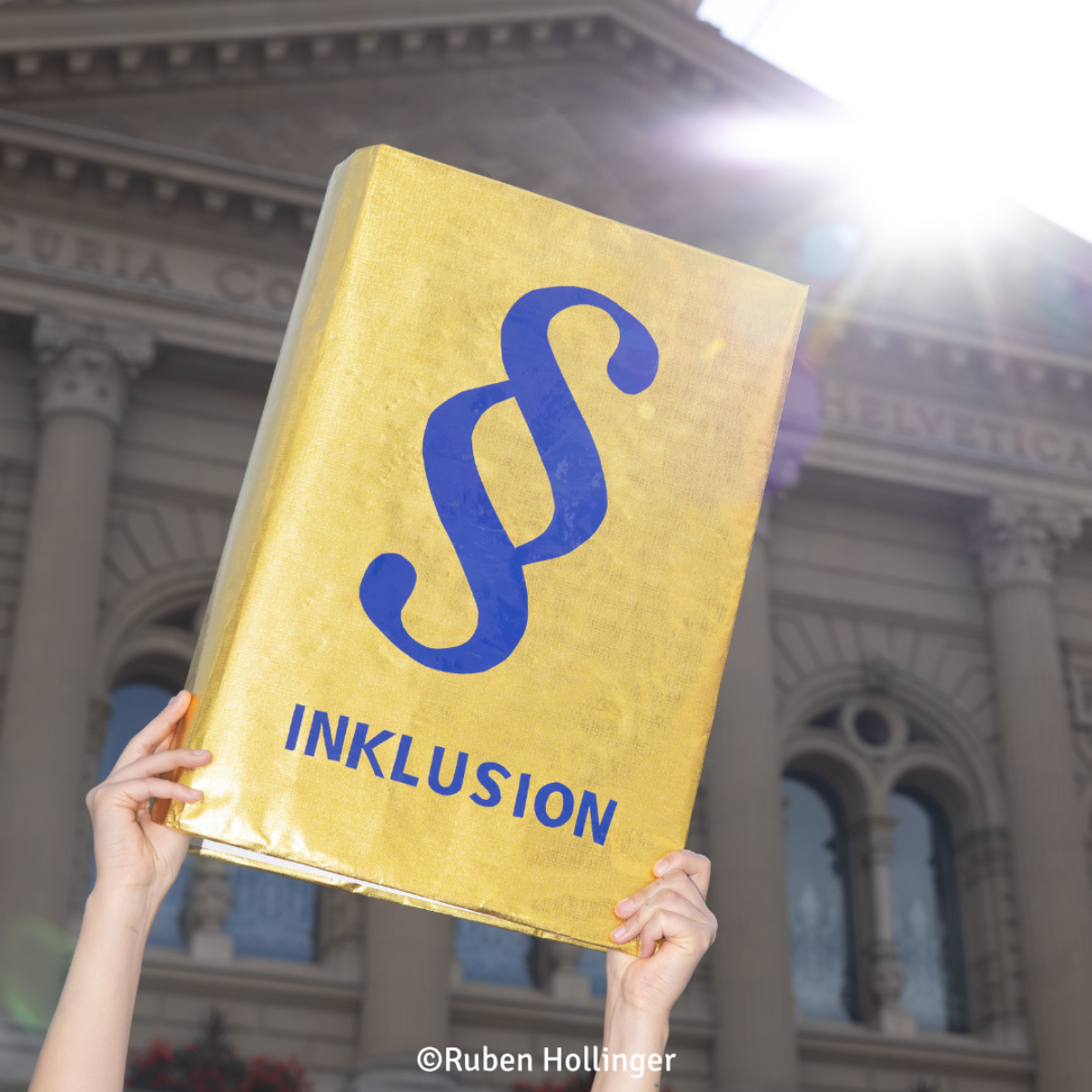 Ein goldener Ordner mit einem Paragraphenzeichen und dem Wort "Inklusion" wird in die Höhe gehalten. Foto von Ruben Hollinger