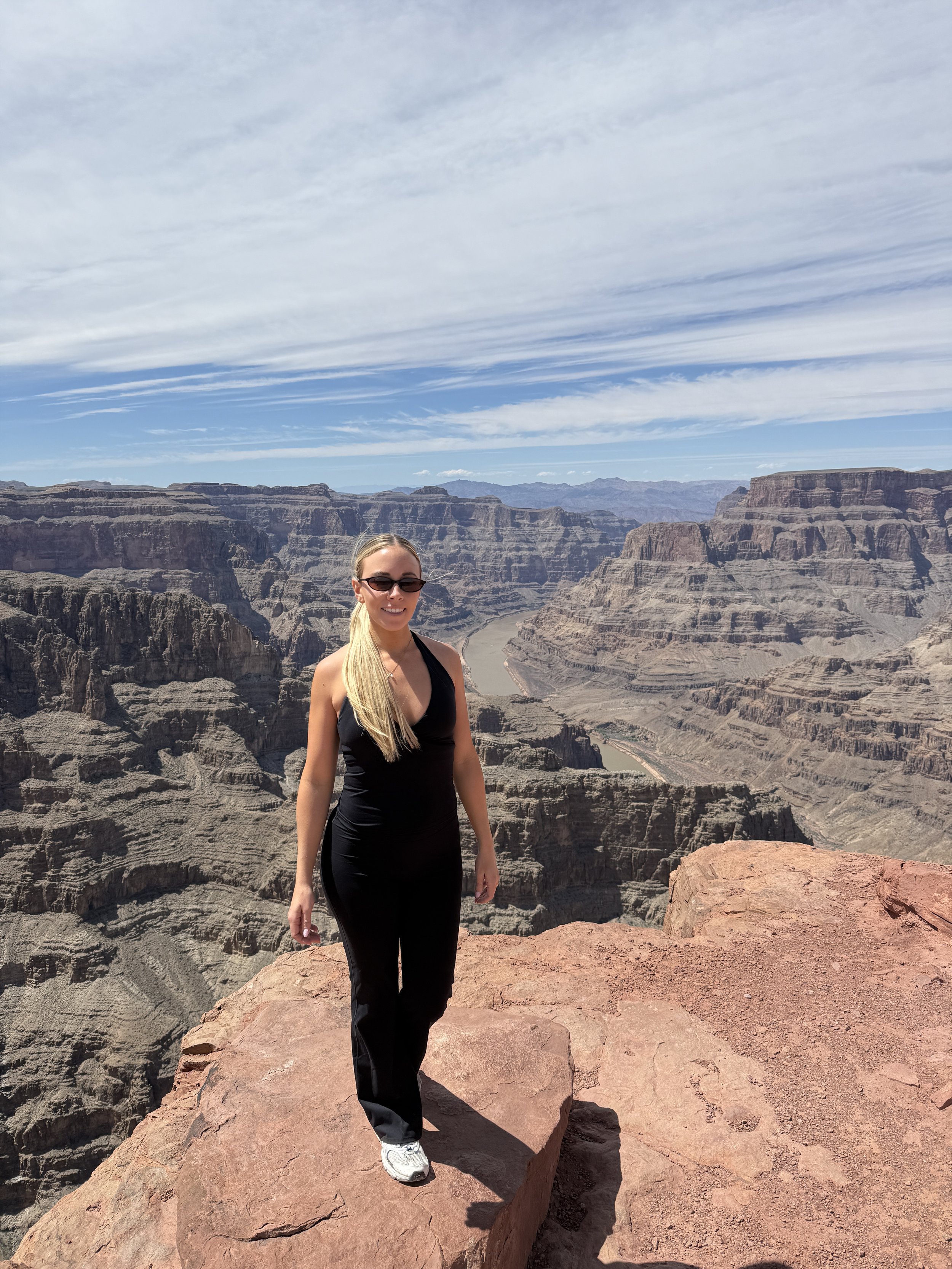 Woman with long blonde hair, wearing sunglasses, a black sleeveless top, and black pants, standing on a large rock at the Grand Canyon with a view of the canyon's cliffs and the Colorado River below, under a mostly cloudy sky.