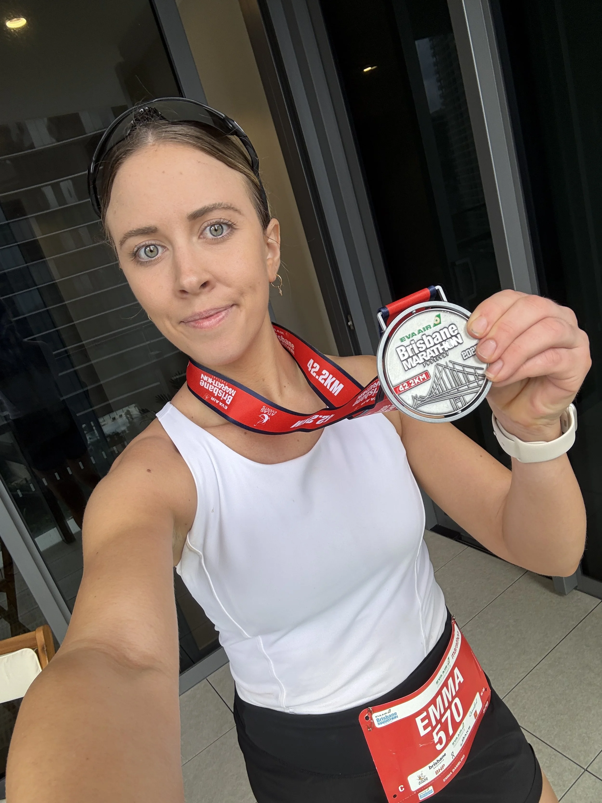 A woman with blue eyes and light brown hair holding a finisher medal from the Brisbane Marathon, wearing a running bib with the number 570 and event details, dressed in a white tank top and black shorts, standing outdoors.