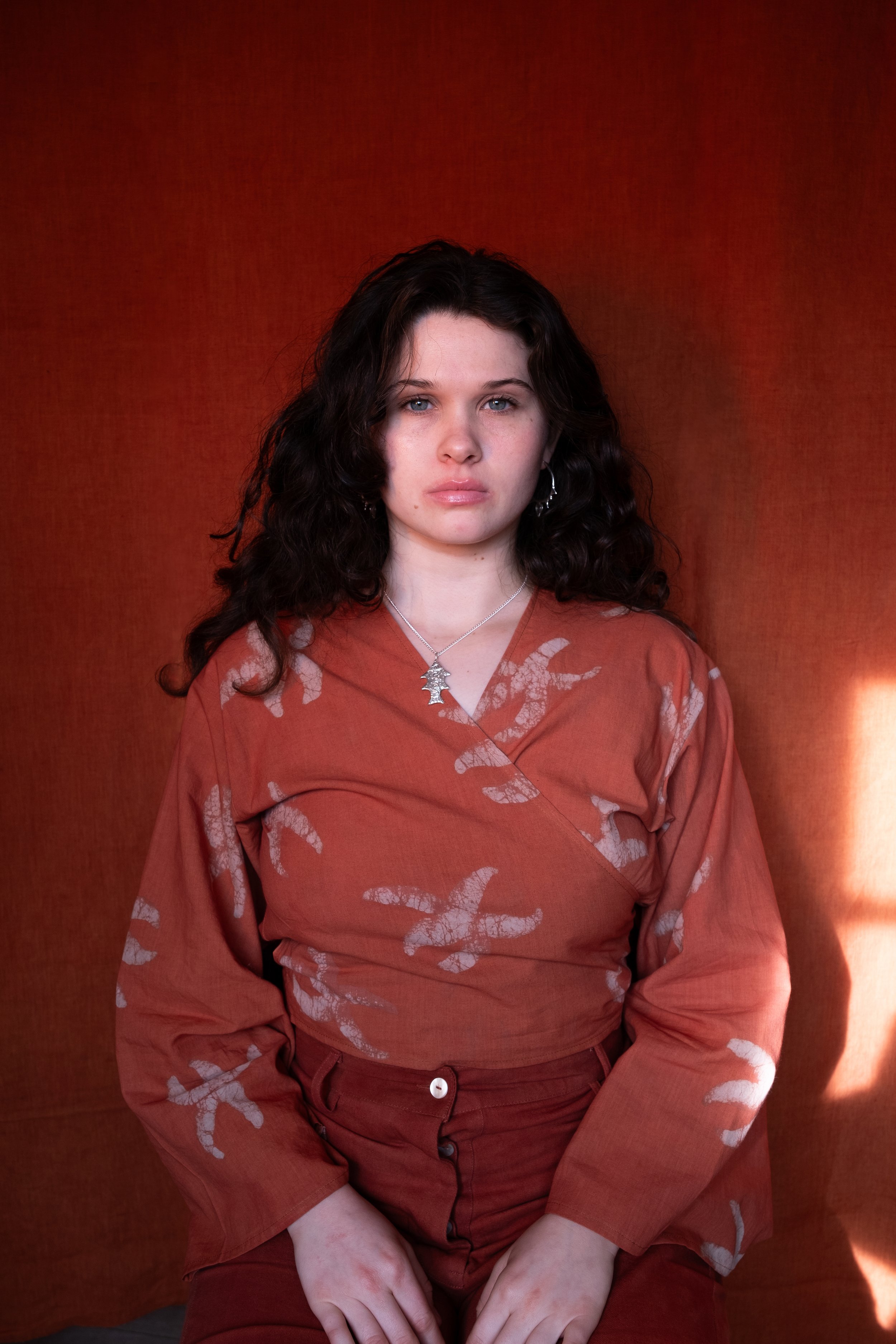 Woman with dark hair sat in front of a dark orange back drop wearing our Yema wrap top in clementine with stafish pattern