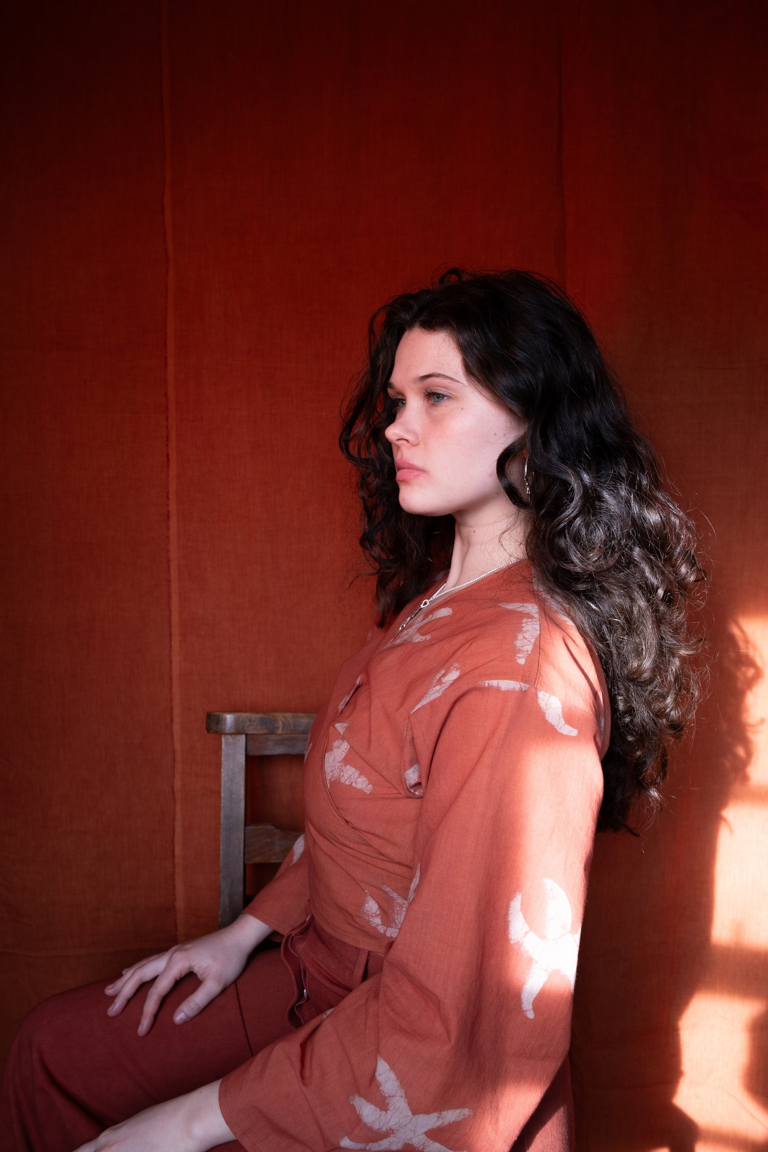 Side on shot of a woman with dark hair sat in front of a dark orange back drop wearing our Yema wrap top in clementine with stafish pattern