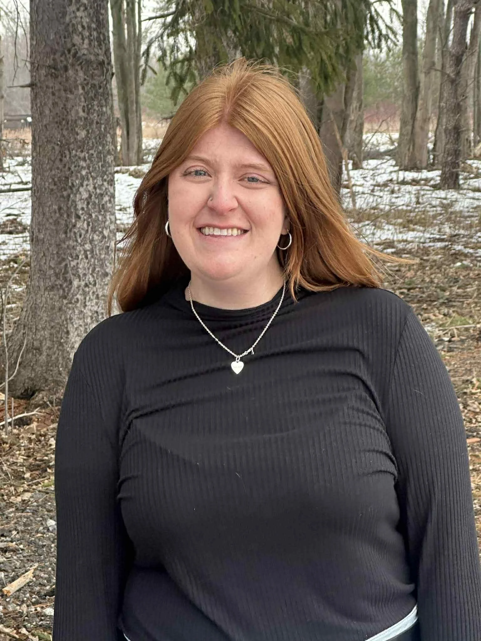 A woman with red hair and fair skin smiling outdoors in a wooded area with snow on the ground, wearing a black ribbed long-sleeve shirt, silver necklace with heart pendant, and silver hoop earrings.