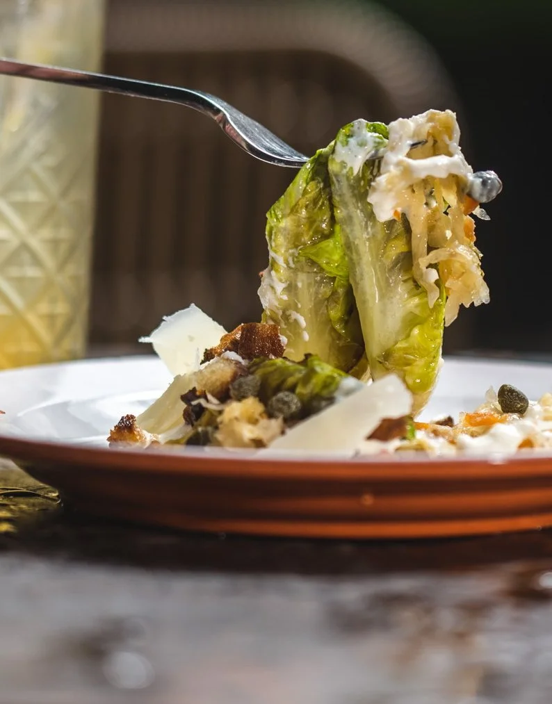 A salad with lettuce, cheese, croutons, and black peppercorns on a plate, served with a fork; a glass is visible in the background.