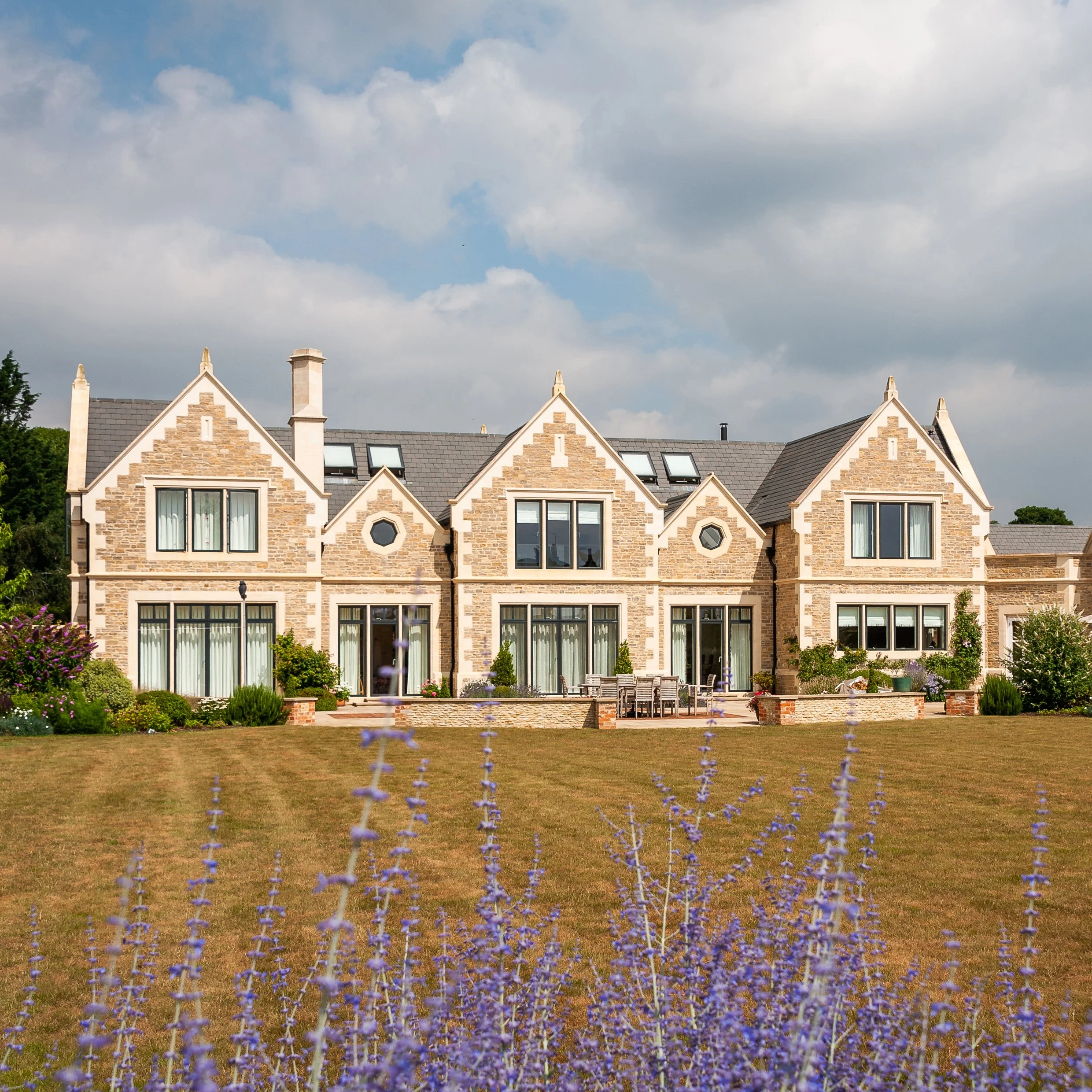 Rear elevation with generous garden space of substantial new home, influenced by Edwardian Arts and crafts architecture and made from natural stone walling, bespoke dressed stone detail and large modern windows.