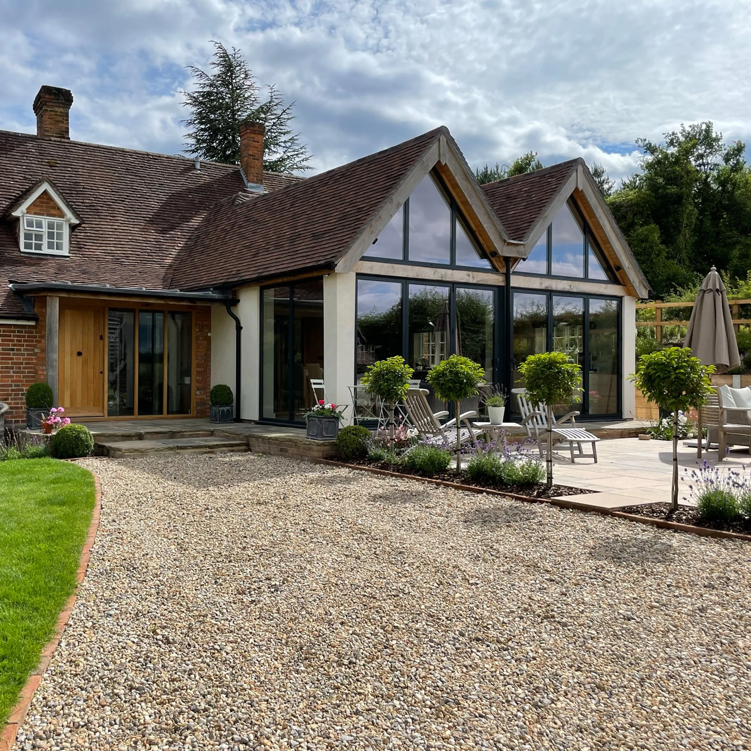 Modern dual-gable glazed ground floor extension to a 17th century house with adjacent outdoor patio and seating, as seen from entrance pathway.
