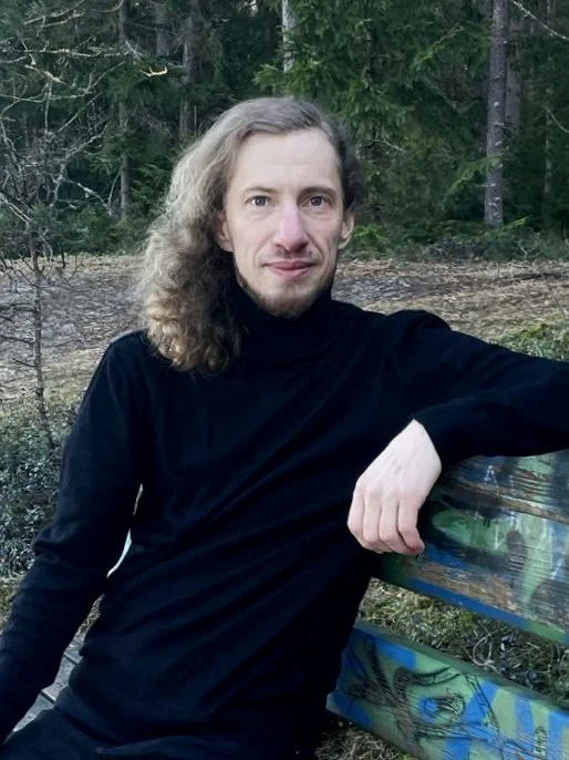 Susi Nousiainen is sitting on a bench, watching straight to the camera and smiling.
