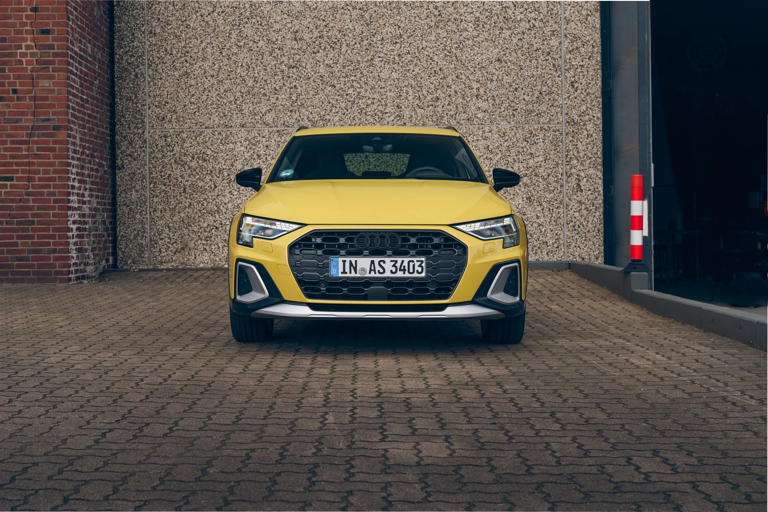 Ein gelber Audi A3 ist geparkt auf einem gepflasterten Parkplatz vor einer Wand mit einer roten Backsteinmauer auf der linken Seite und einer geometrischen Steinwand im Hintergrund.