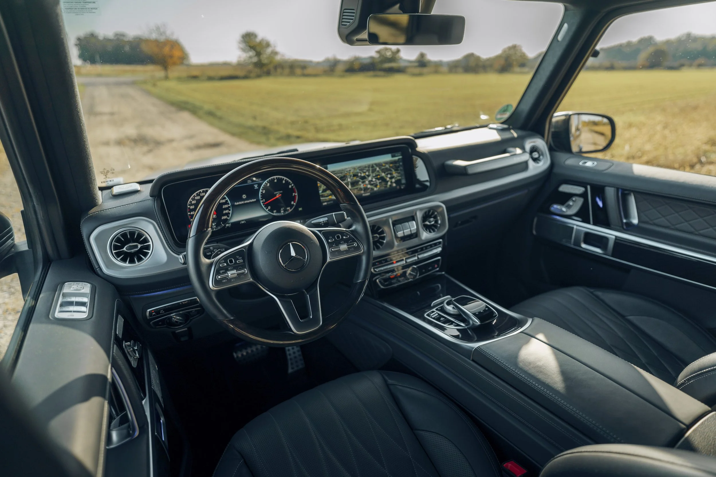 Innenansicht eines Mercedes-Fahrzeugs mit Digitalanzeigen, Lenkrad, Navigationsbildschirm und schwarzen Ledersitzen, das auf einer Landstraße fährt.
