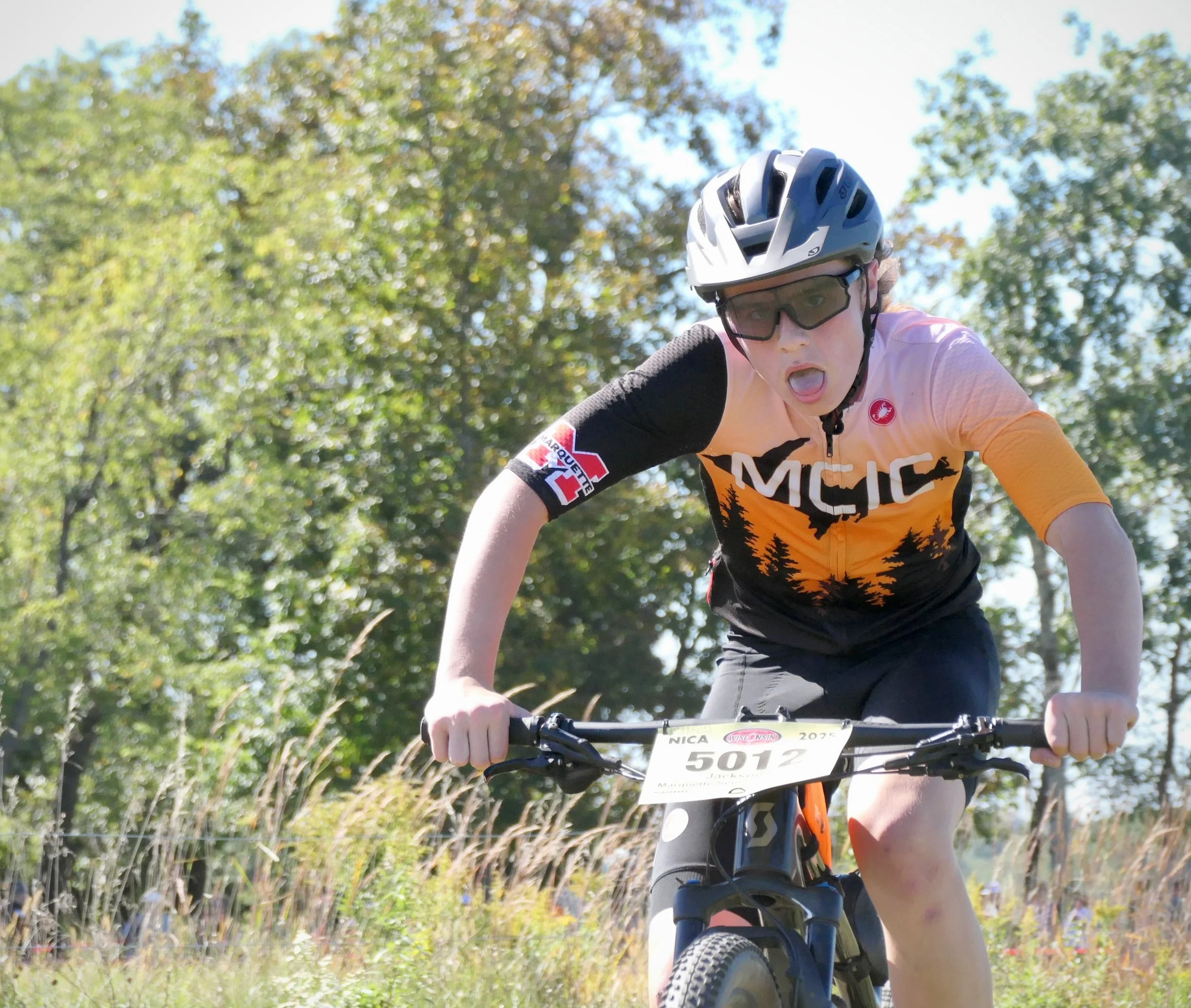 Young person riding a mountain bike on a trail, wearing a helmet and sunglasses, with a race number 5012 attached to the bike, surrounded by trees and grass on a sunny day.