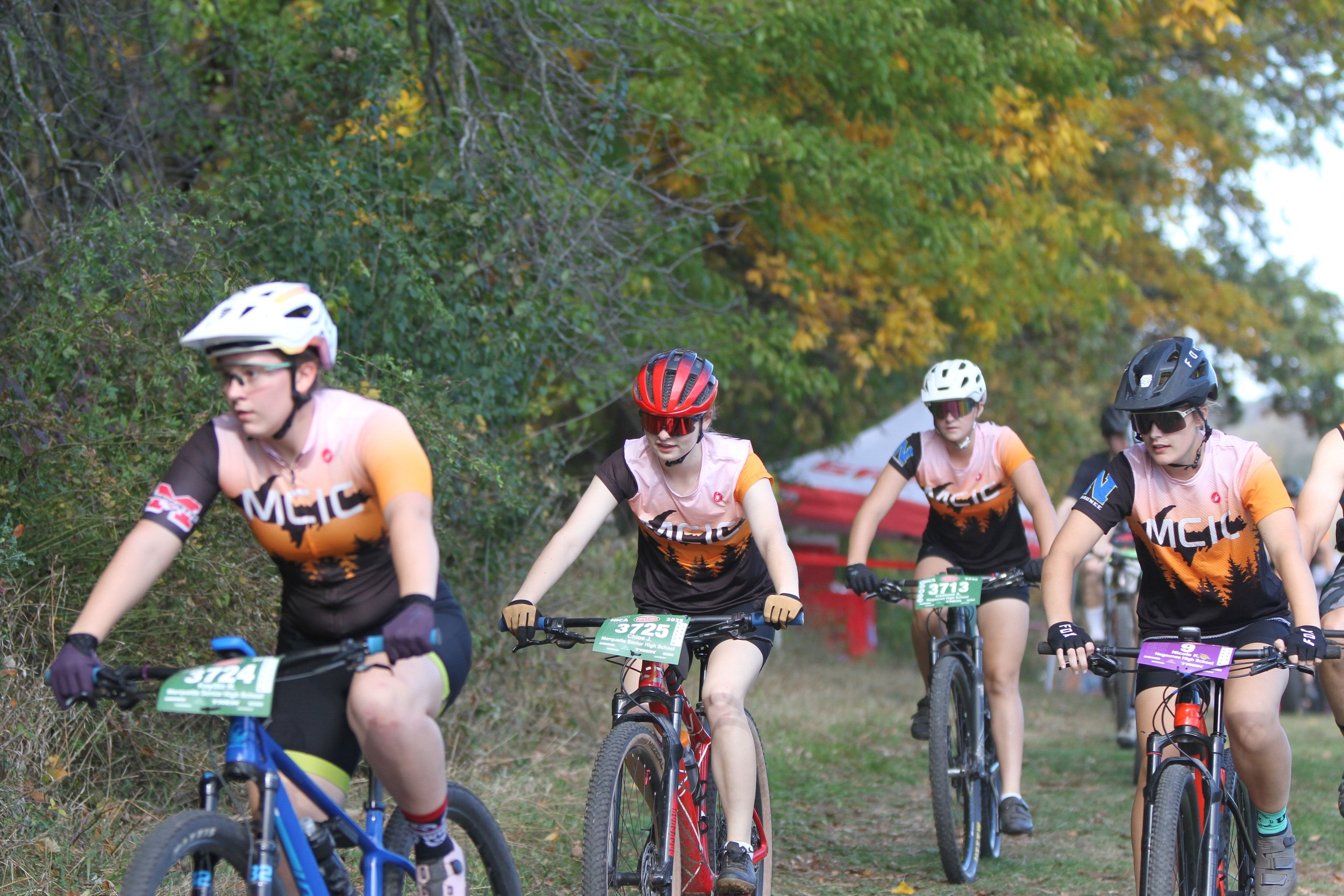 Group of female mountain bikers riding on a trail through a wooded area during daytime.