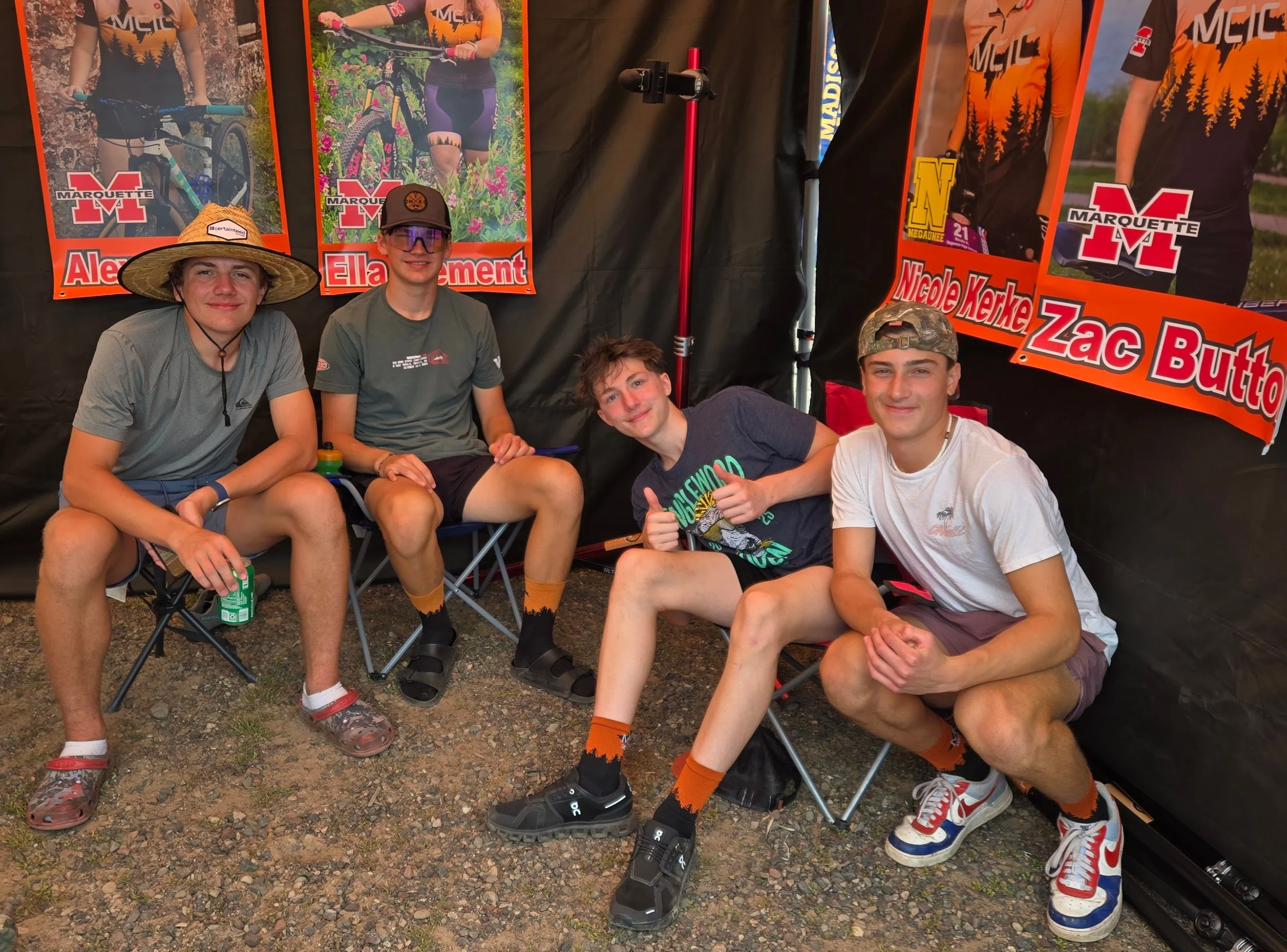 Four young men sitting in front of a Marquette High School cycling team display, with posters of cyclists and their names in the background. They are outdoors, wearing casual clothes, some with cycling shoes and socks, and are posing happily for the 