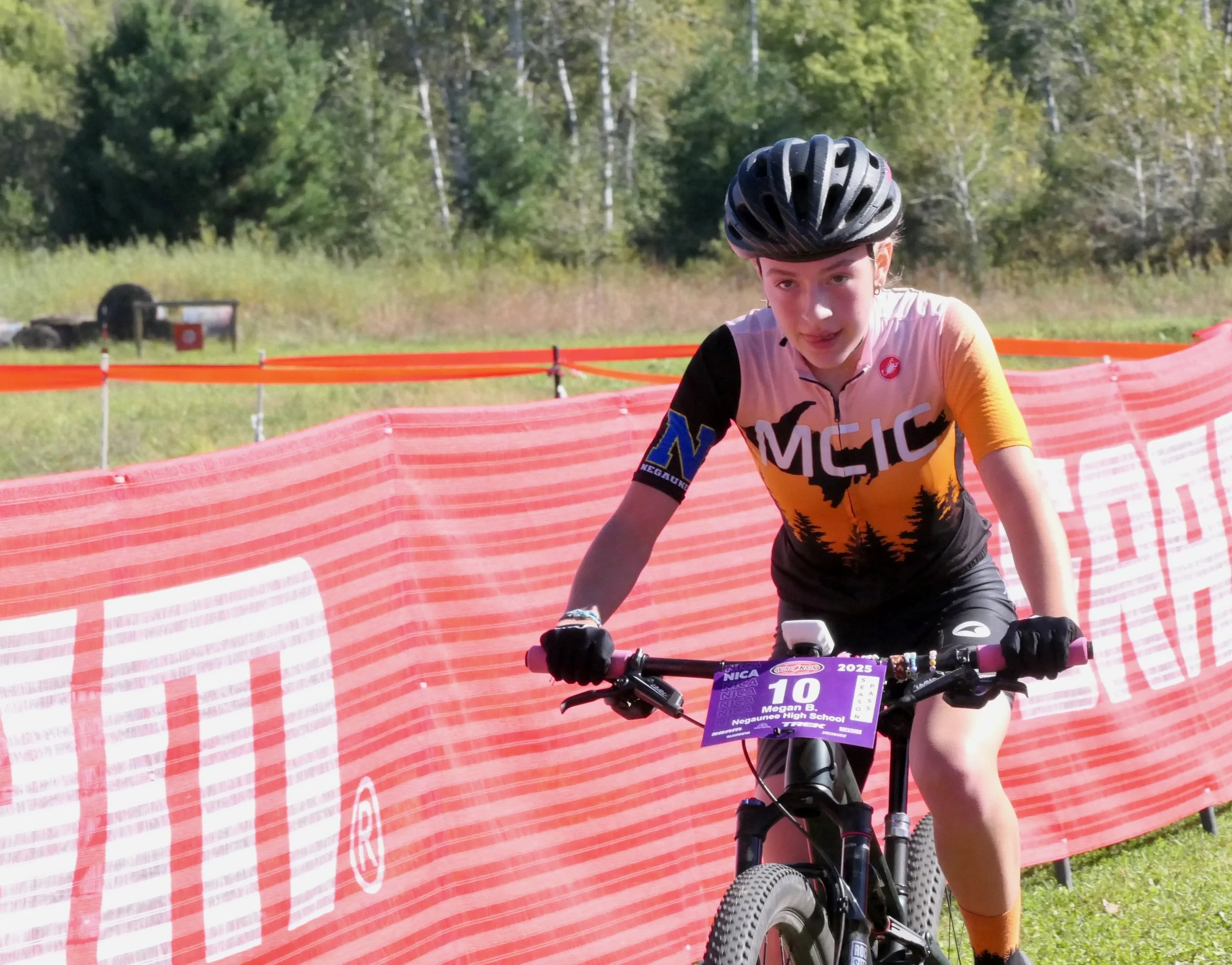 Young female cyclist in racing gear riding her mountain bike on a dirt course, with a red safety barrier and green wooded area in the background.