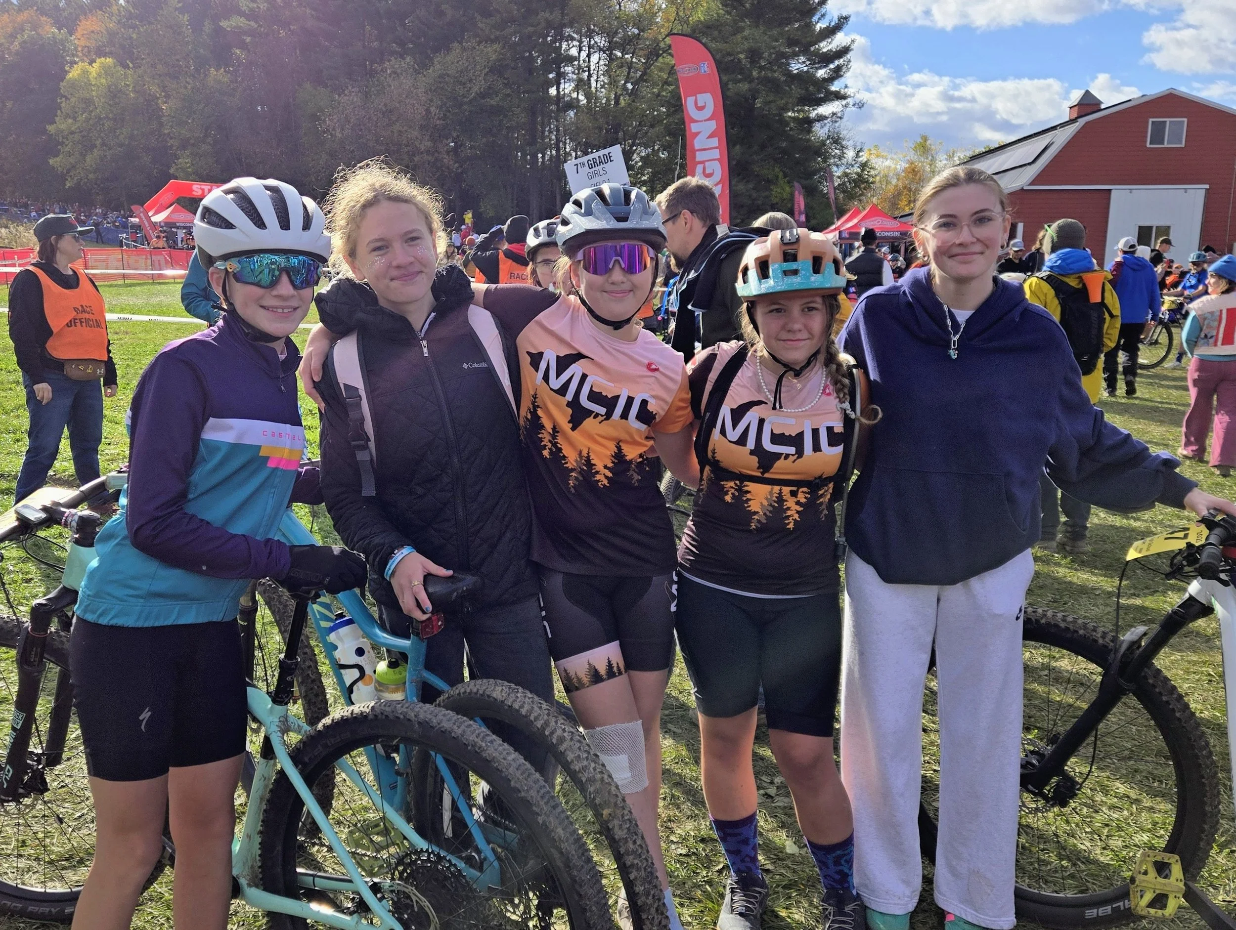 Group of five young girls with bicycles at an outdoor cycling event, wearing helmets and athletic clothing, smiling and posing for the camera with a crowd and banners in the background.