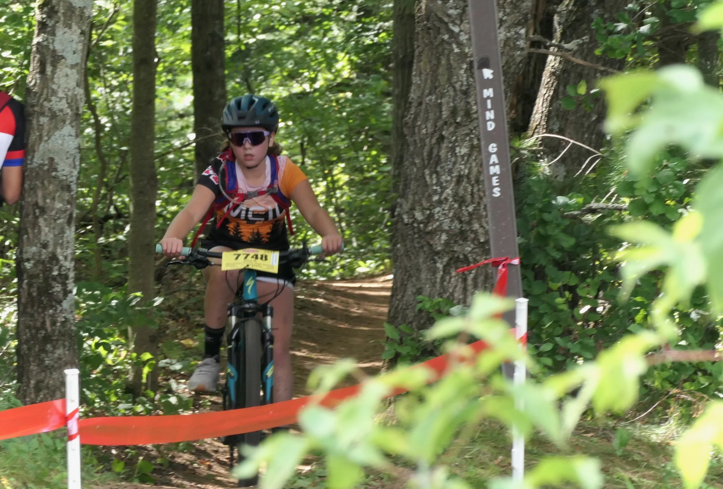 A young girl wearing a helmet and sunglasses riding a mountain bike through a wooded trail during a race or event, with a bib number 7748 visible, surrounded by trees and green foliage.