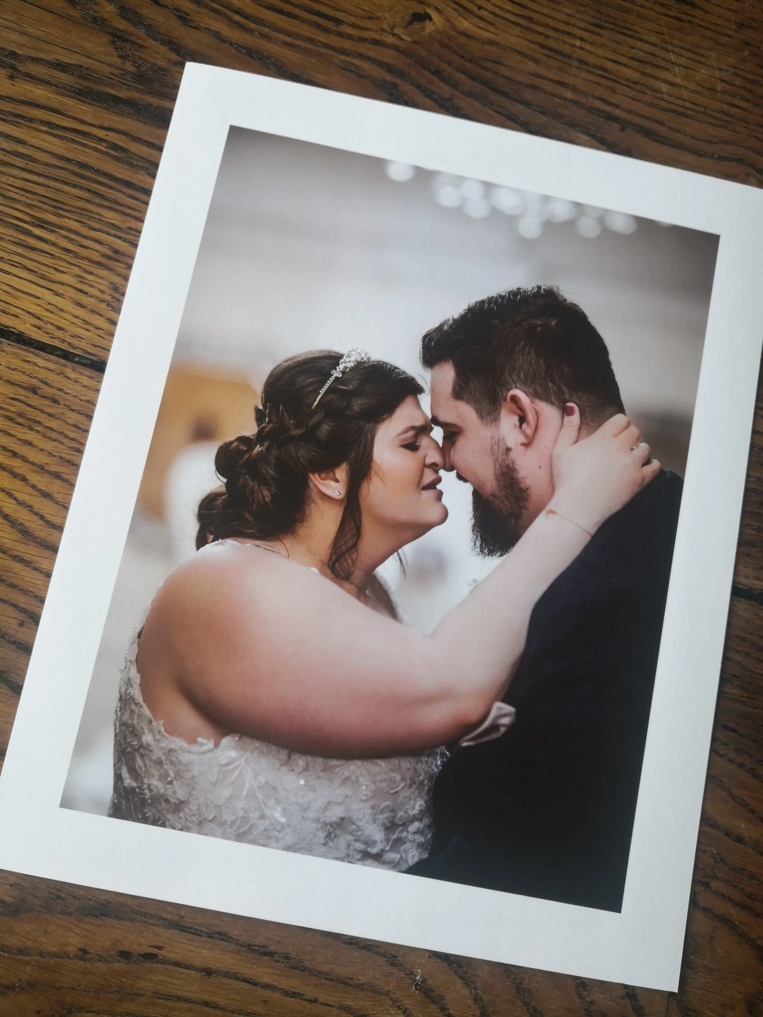 A wedding photo of a bride and groom touching foreheads with their eyes closed, the bride has dark wavy hair and is wearing a lace wedding dress, the groom has short dark hair and a beard, both are dressed in wedding attire.