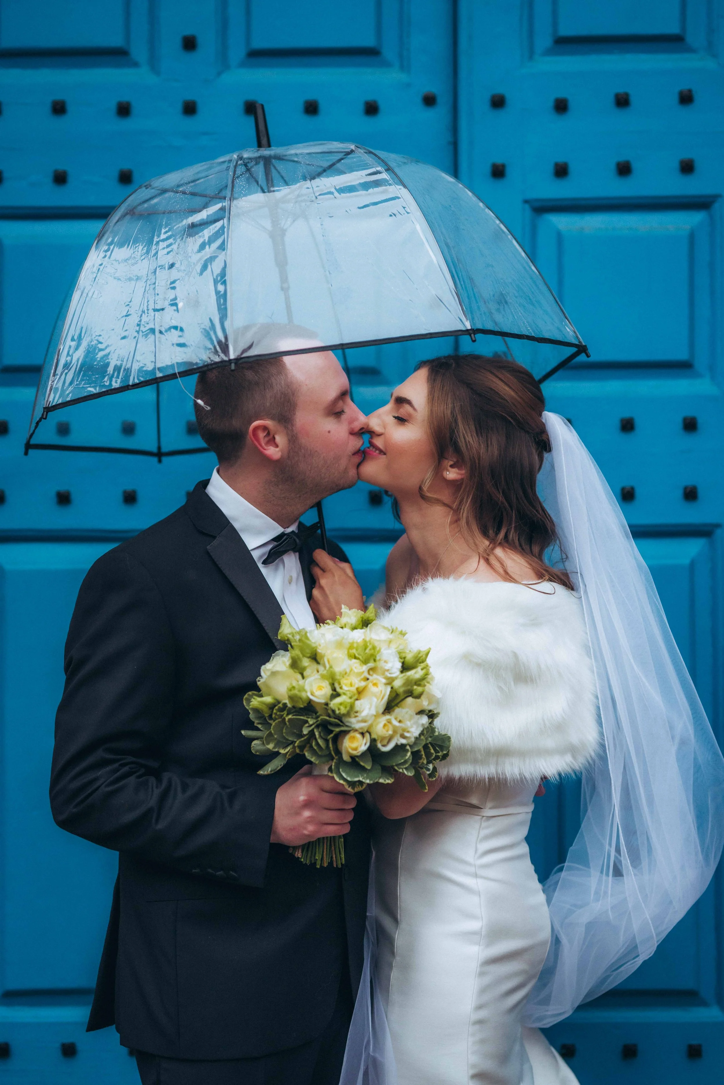 Bride and groom kissing under an umbrella and in front of a blue wooden gate at Gosfield Hall in Halstead