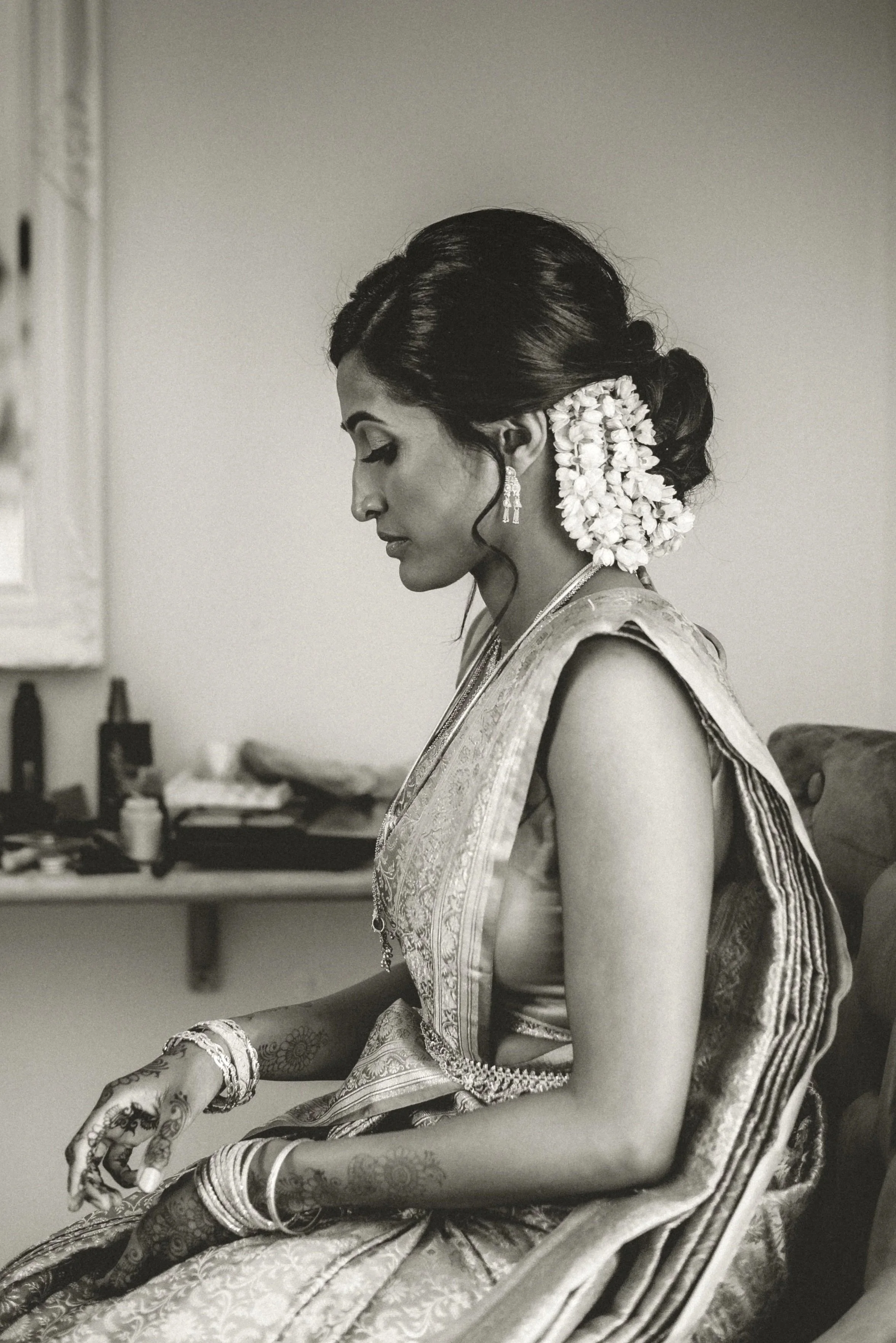 A bride in a traditional wedding dress and flowers in her hair is sitting on a chair looking down to her hands covered in henna tatoos at East Mersea Hall near Colchester