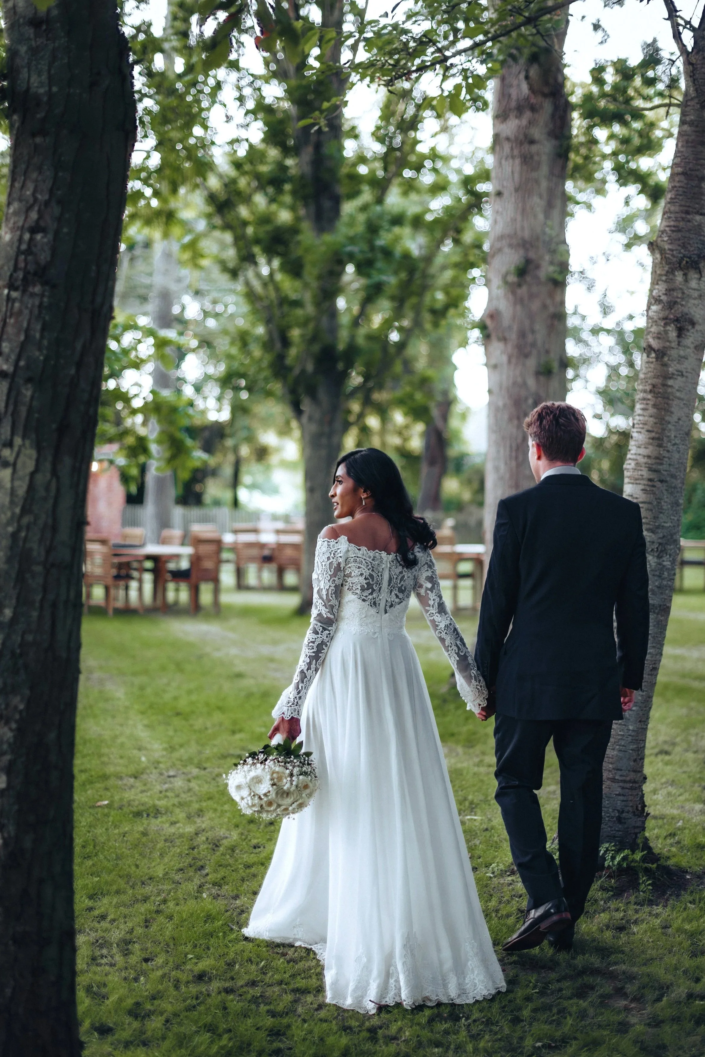 Bride and groom are holding hands and walking through an area of trees and lawn at East Mersea Hall in Colchester