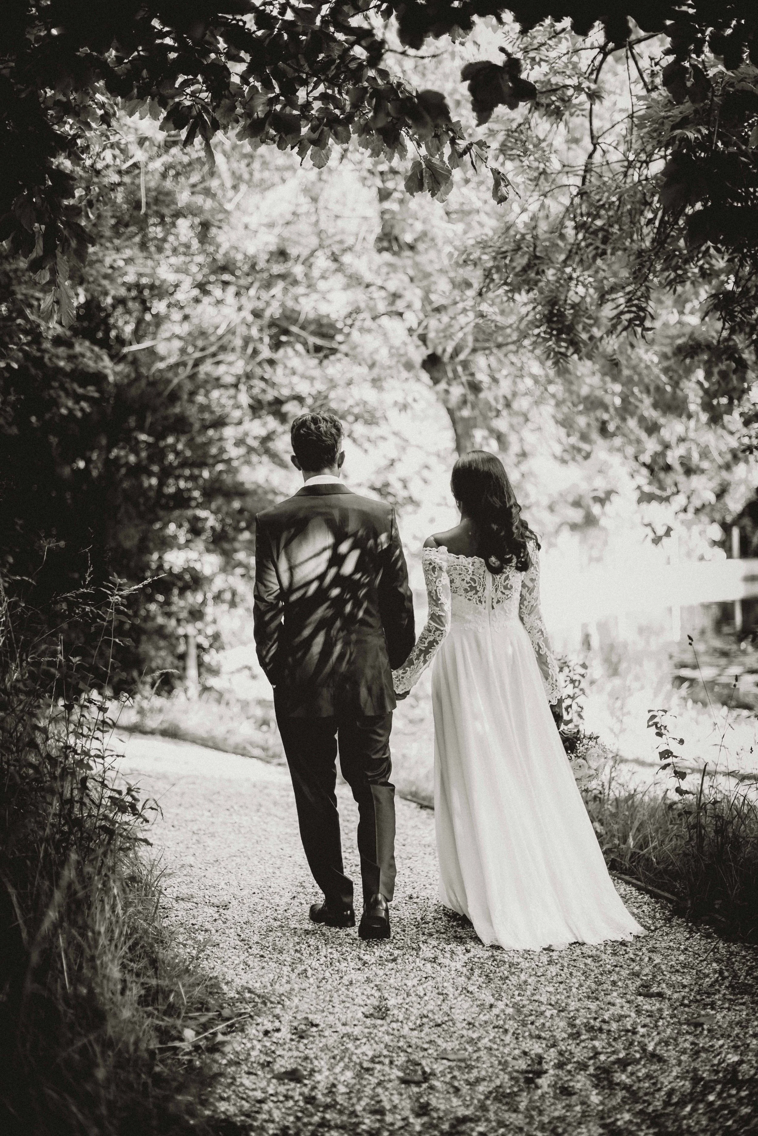 A black and white photograph of a bride and groom walking hand in hand down a garden path surrounded by trees and foliage, with the bride wearing a long lace dress and the groom in a suit.