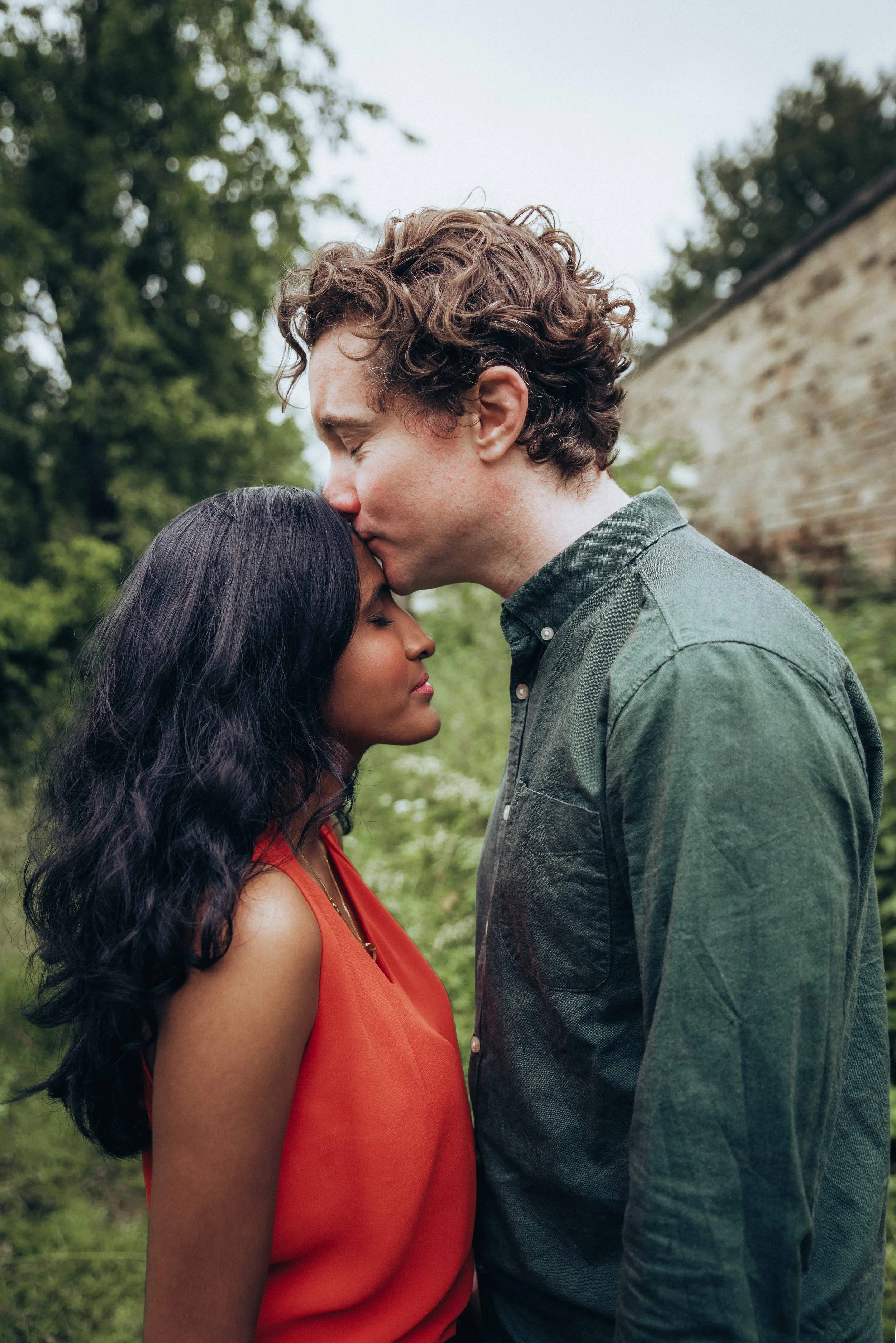 A couple standing and facing each other while he kisses her forehead during an engagement session in a park near Cambridge in summer