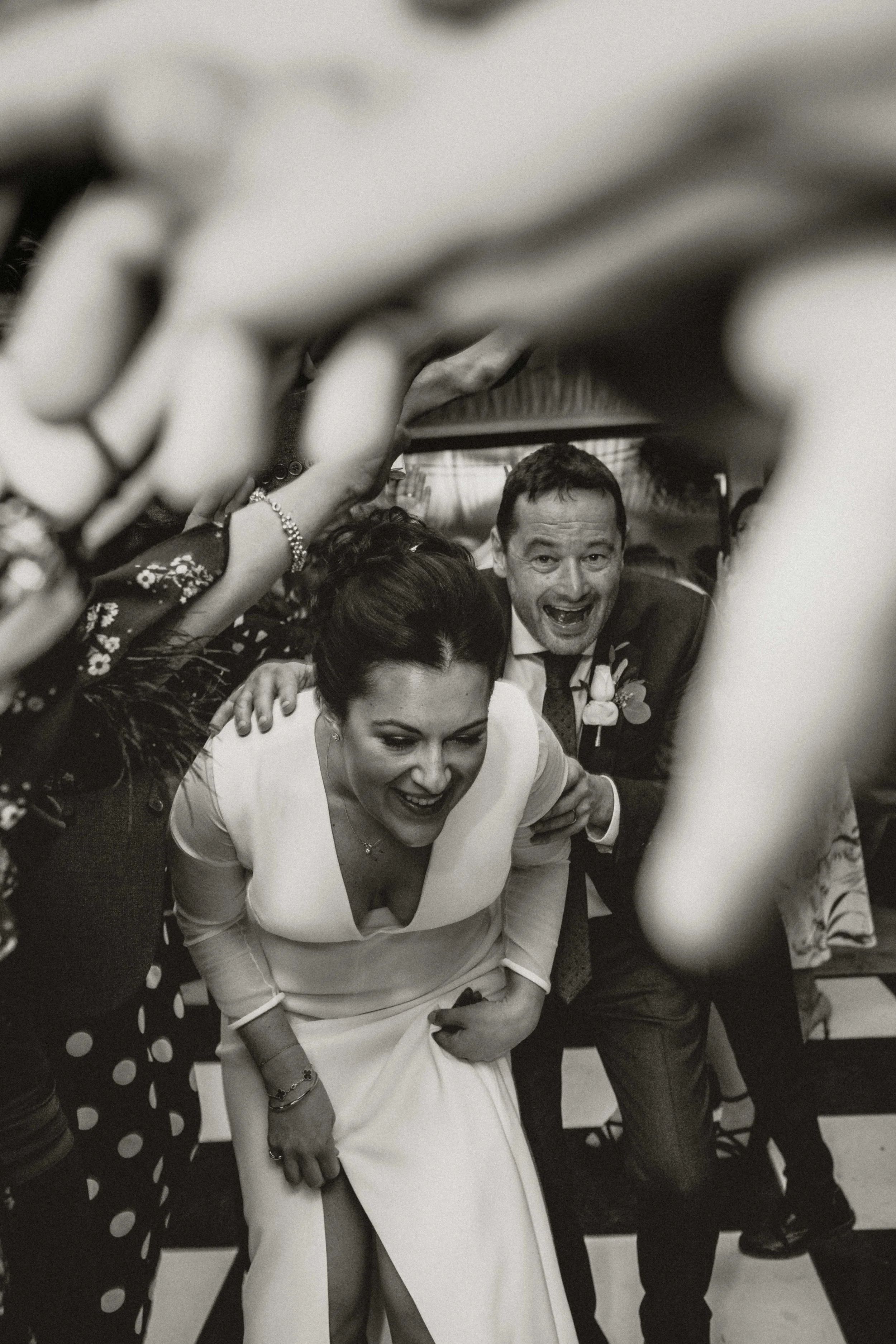 A black-and-white photo of a group of people at a celebration or party. A woman in a white dress is in the center, smiling and leaning forward. Other guests are around her, some reaching out or touching her. The scene appears lively and joyful.