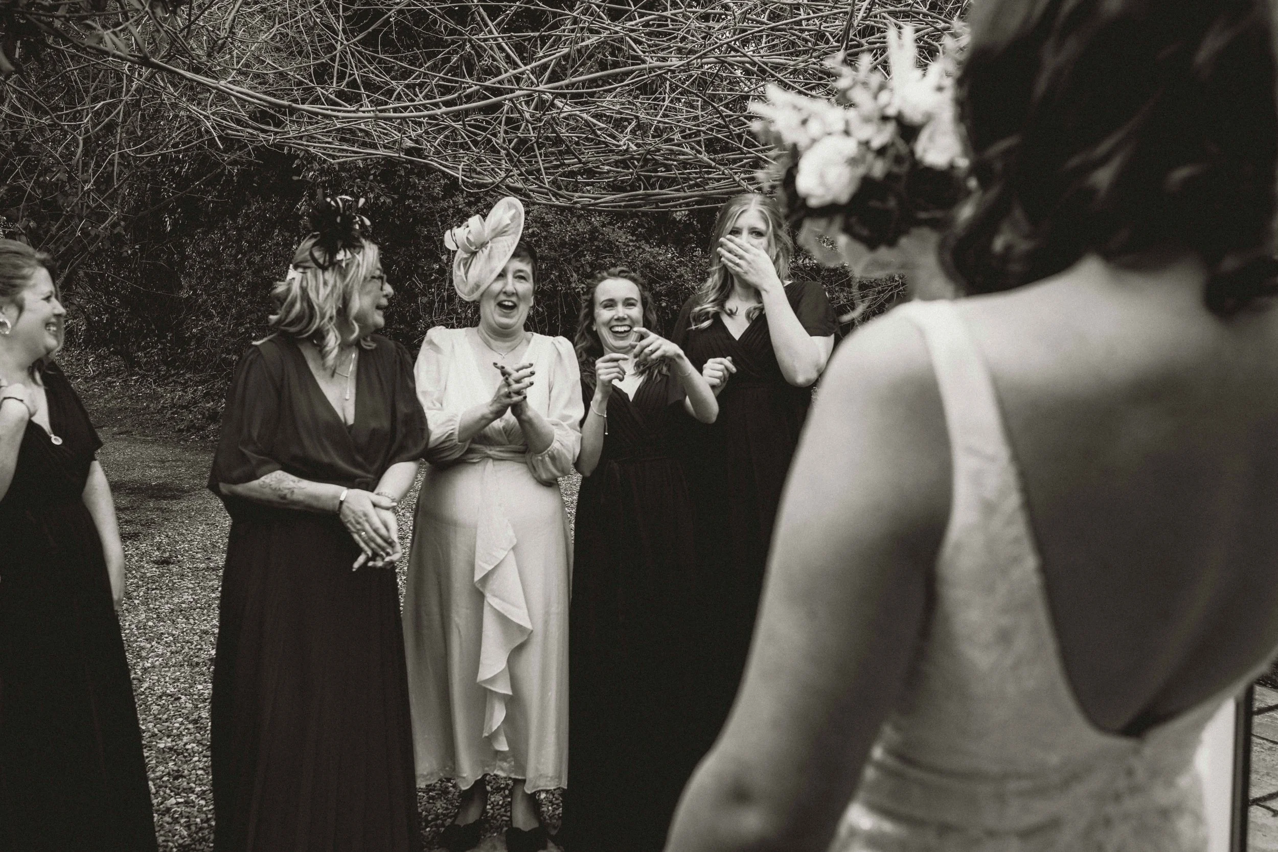 A group of women standing outdoors, dressed in formal attire, laughing and smiling during a gathering or celebration. One woman covers her mouth as she laughs, wearing a hat, and the others appear to be enjoying the moment in front of a backdrop of l