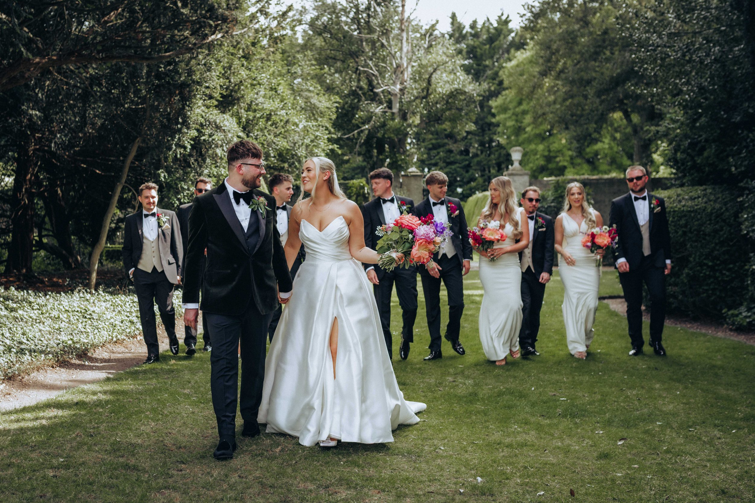 A bride and groom walking hand in hand with a wedding party in the gardens of Hockwold Hall near Brandon in Norfolk.