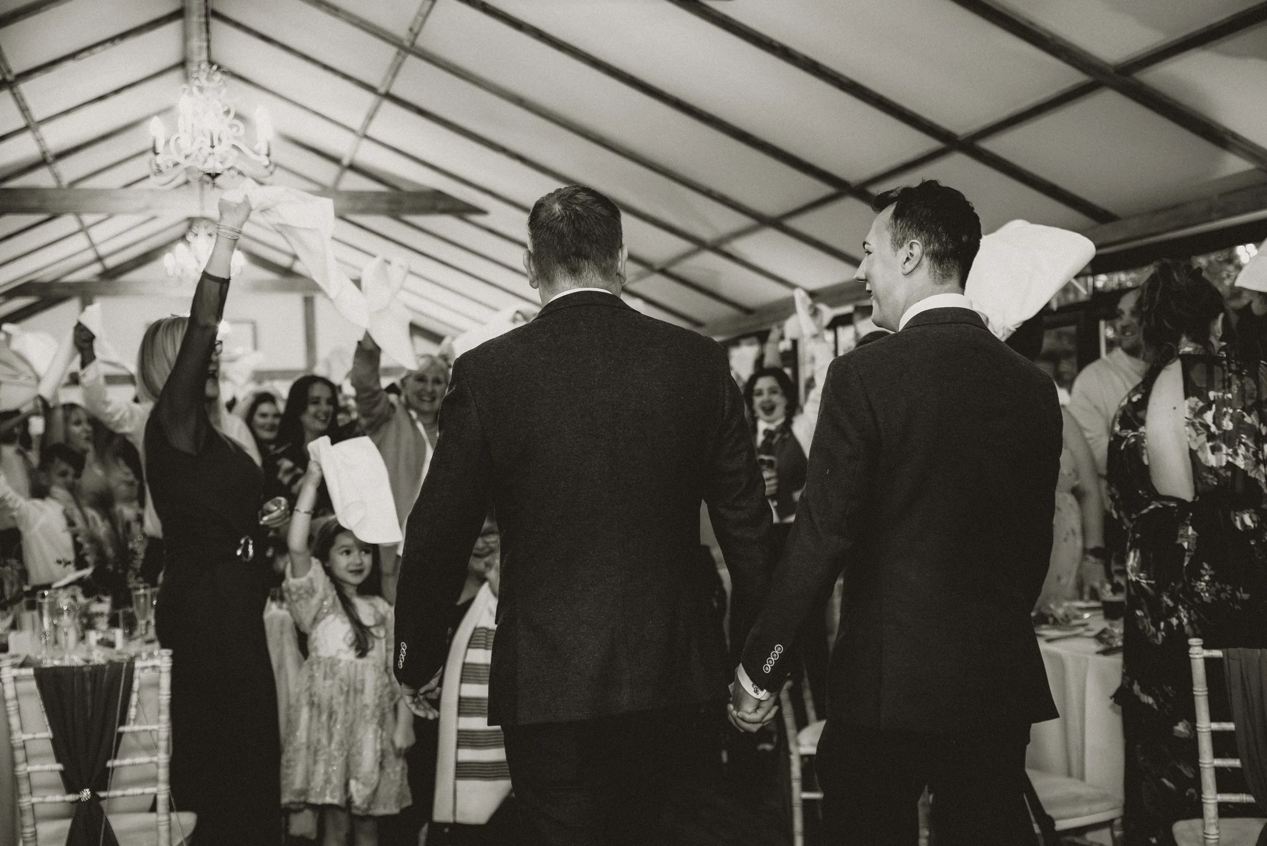 Two men in suits holding hands and facing an audience of smiling women and children during a celebration event in a decorated indoor venue.