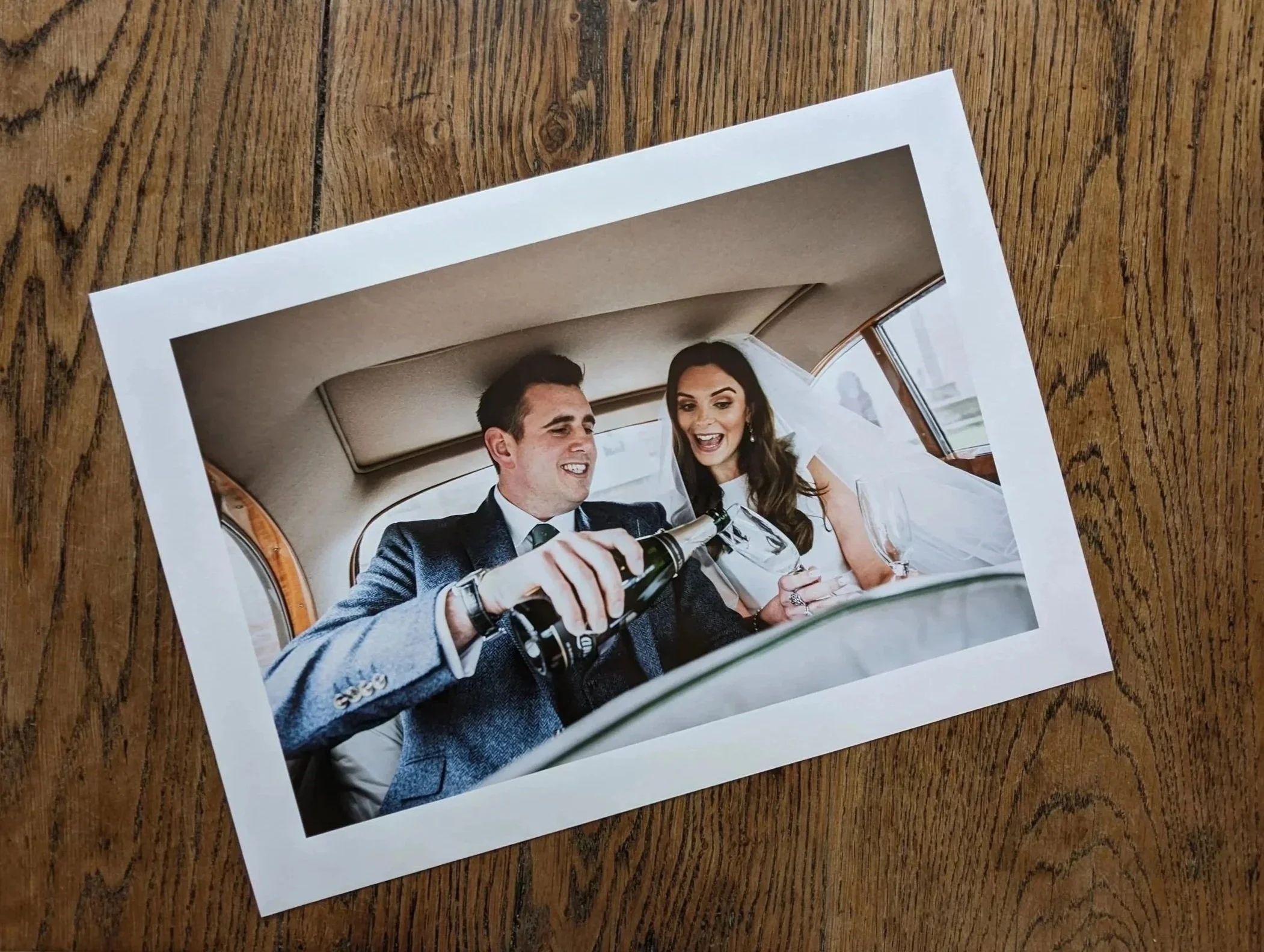 Printed photo of a man and a woman sitting in a car, dressed formally, with the man pouring champagne into a glass held by the woman.