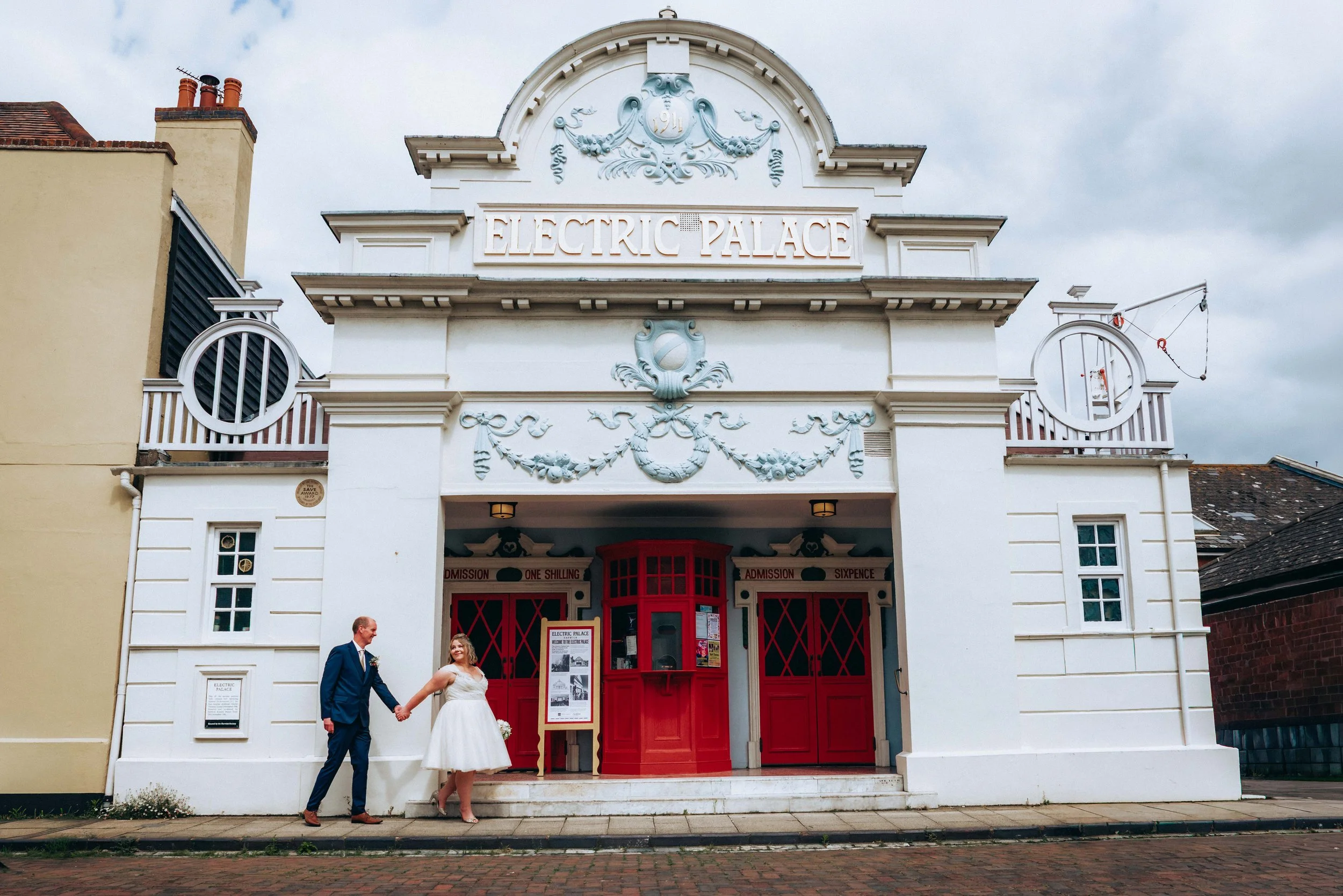 A bride and groom holding hands in front of a vintage-style theater called Electric Palace in Harwich in Essex. The theater has a white facade with decorative details, red doors, and a small ticket booth.