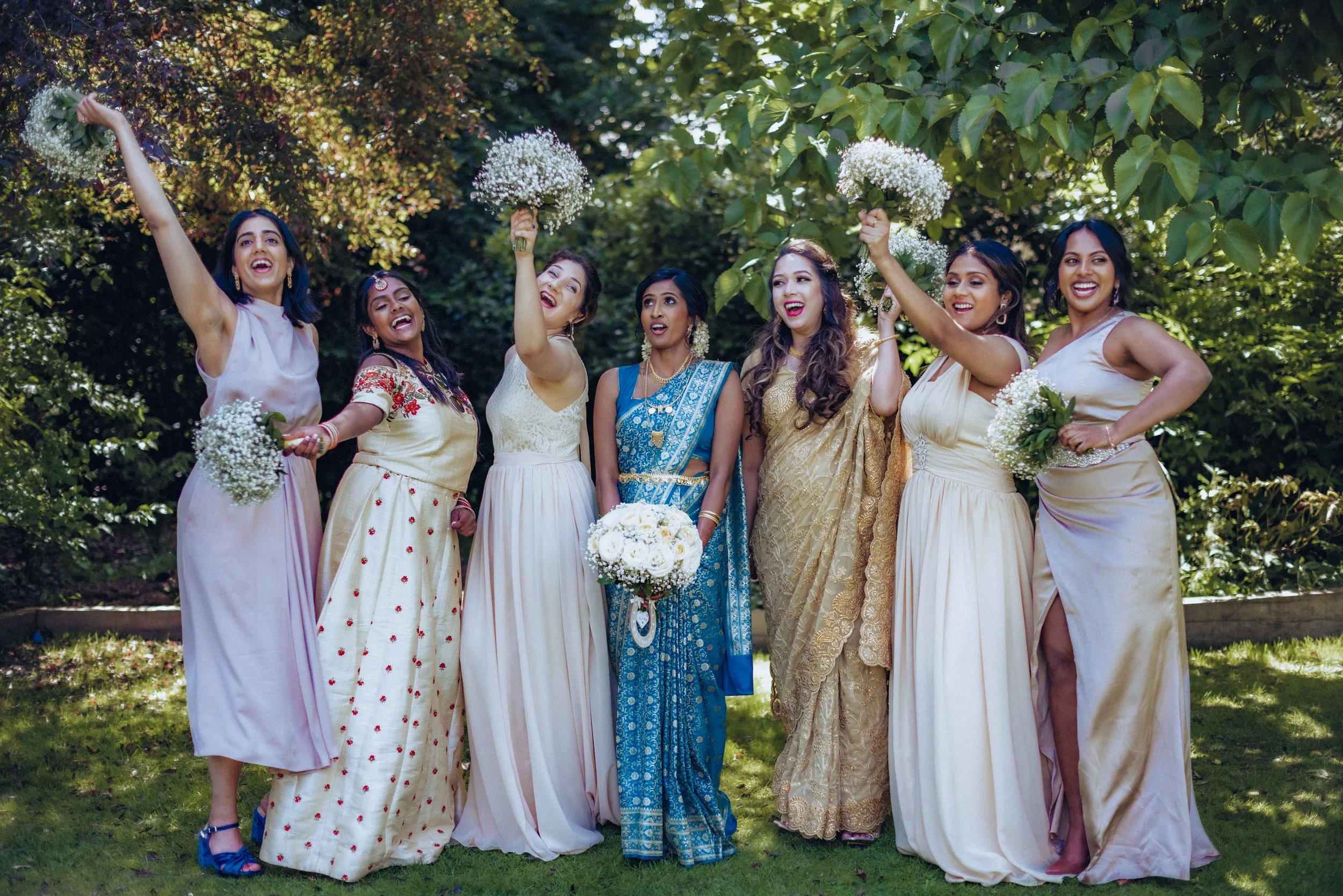 A group of seven women dressed in traditional colorful sari and dresses, smiling and celebrating outdoors in a green park, holding bouquets of white flowers.