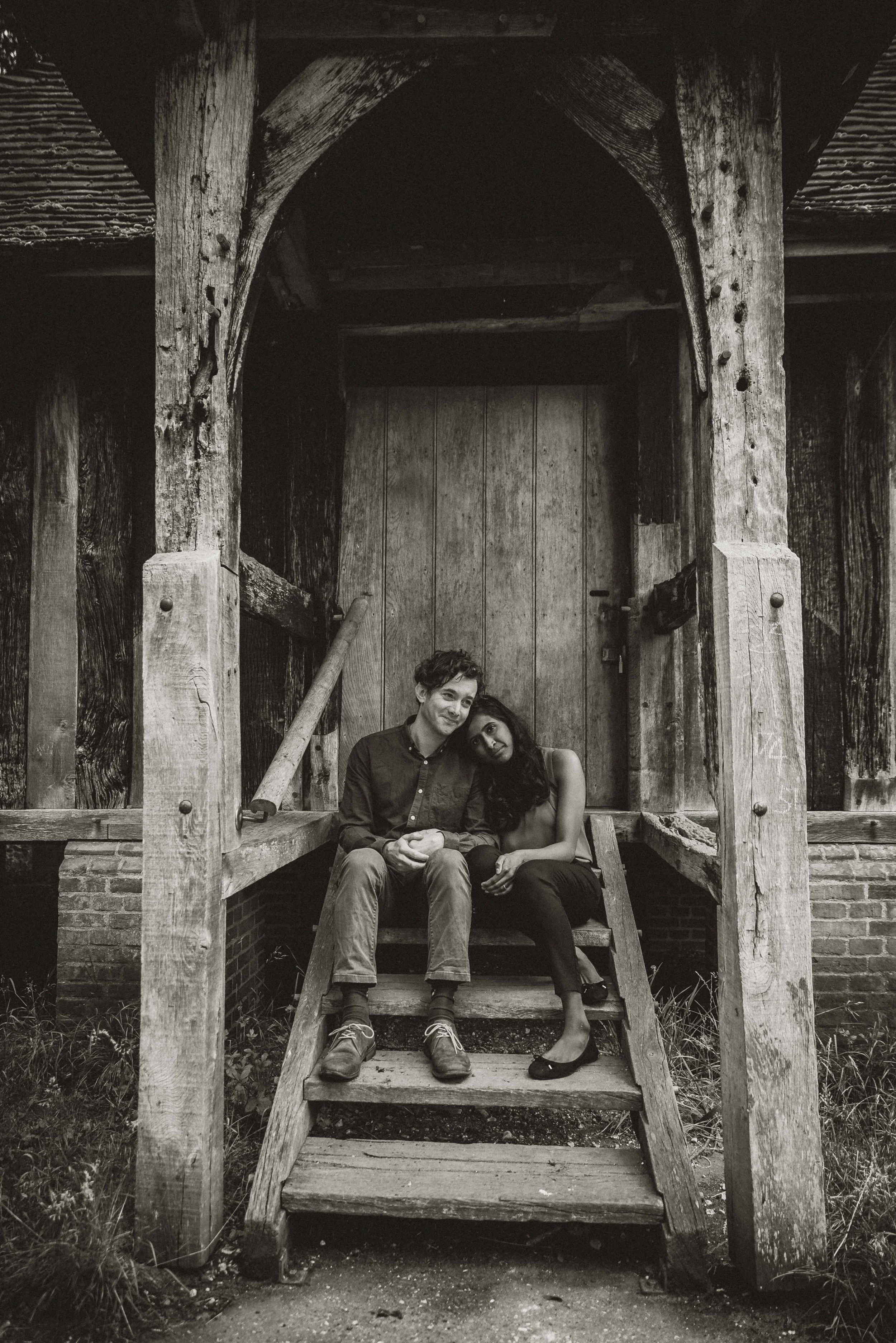 A couple sitting on an old wooden step with her resting her head on his shoulder during an engagement photo session in a park near Cambridge