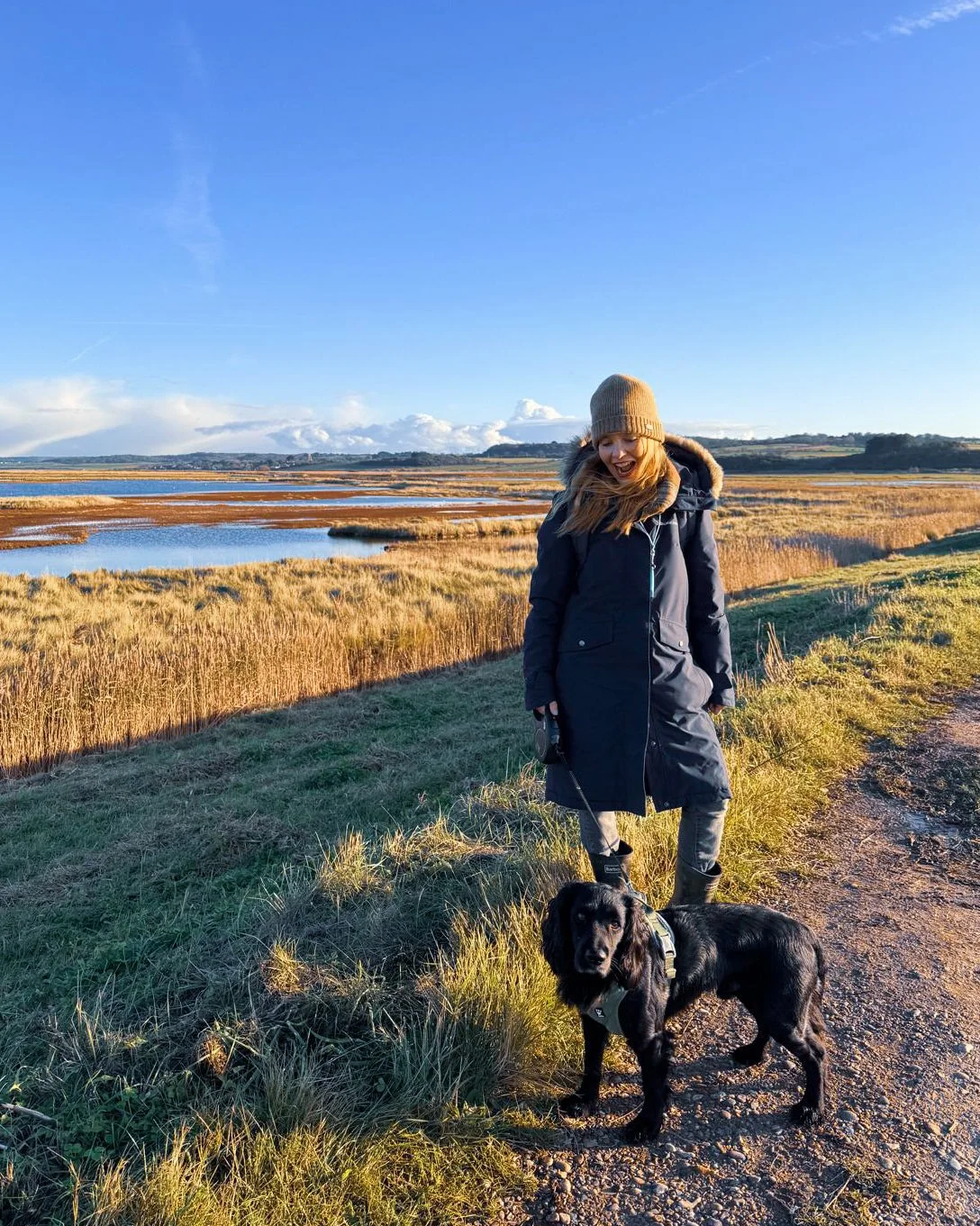 A woman in a navy coat and beige knit hat walking a black dog on a leash in a grassy lakeside area during late afternoon with a clear blue sky.