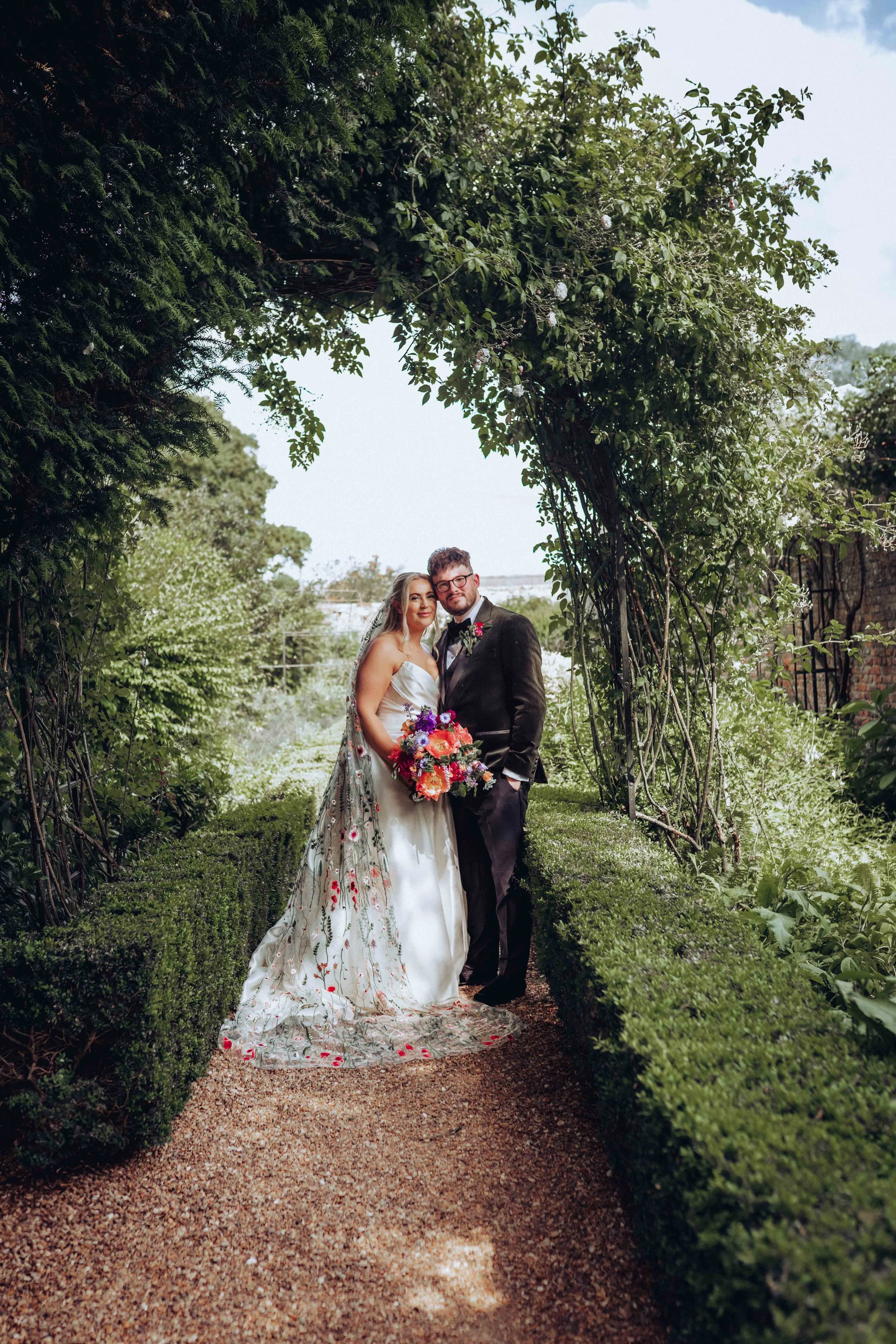 A bride and groom smiling and standing close next to each other underneath a rose arch in the gardens of Hockwold Hall in Hockwold cum Wilton near Brandon in Norfolk