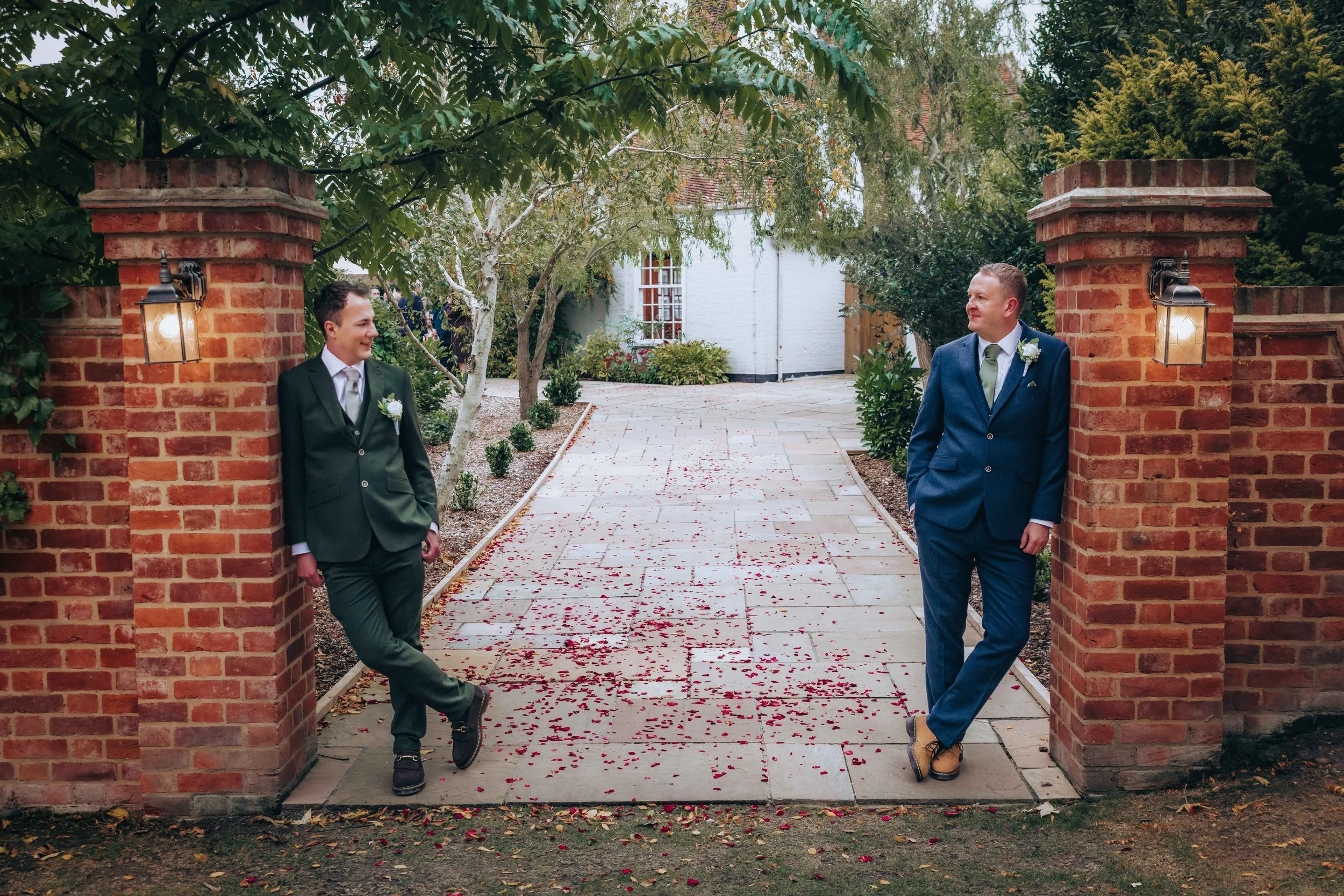 Two men in suits standing on either side of a brick entrance gate, with greenery and a white house in the background, and rose petals scattered on the pathway.