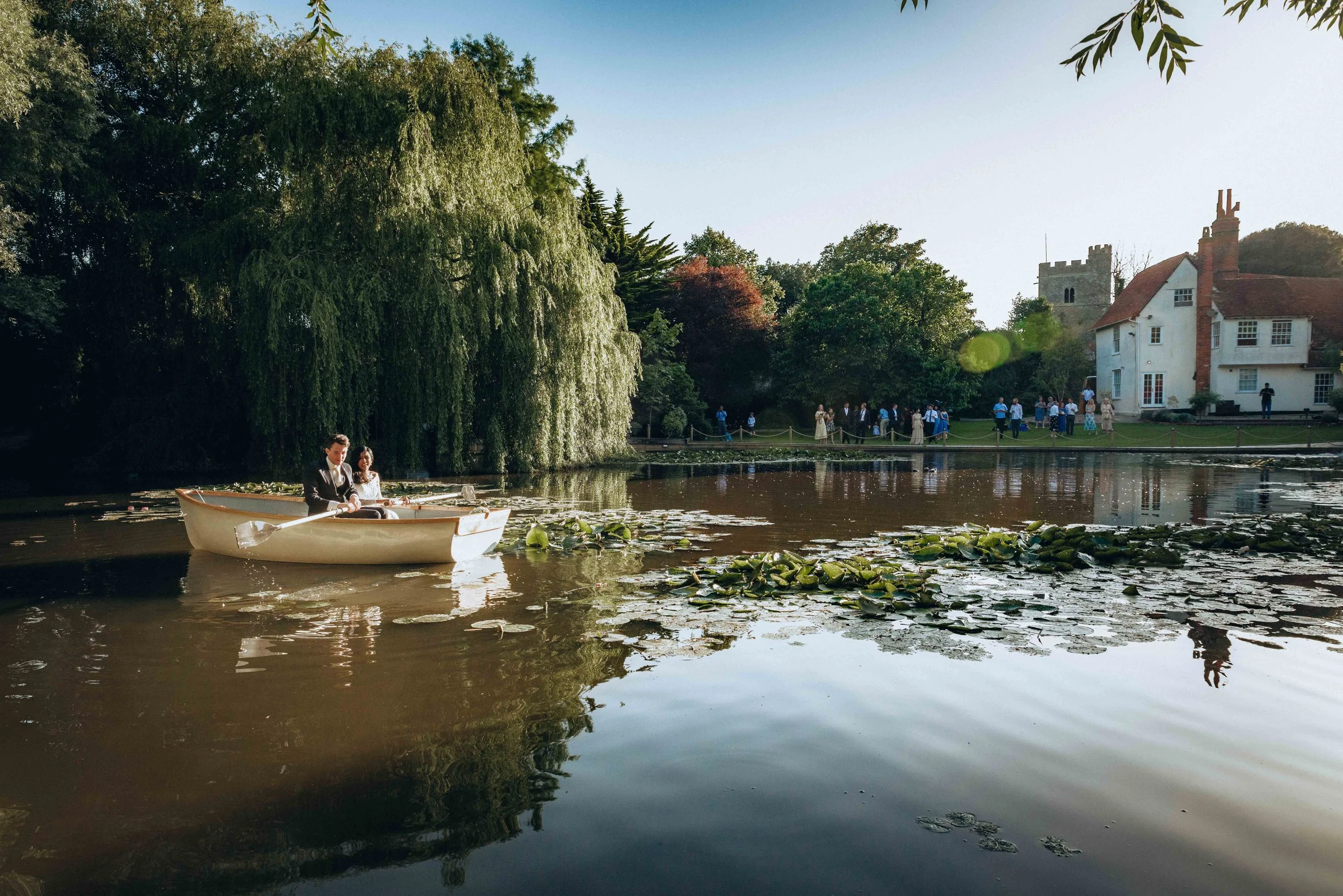 A couple in formal attire sitting in a rowboat on a pond with lily pads, surrounded by lush green trees and a large white house with a tower in the background, under a clear blue sky.