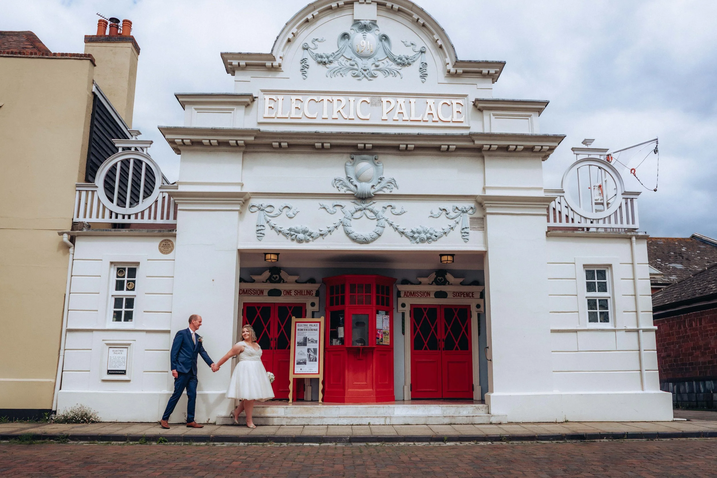 A bride and groom holding hands in front of a vintage-style theater called Electric Palace in Harwich in Essex. The theater has a white facade with decorative details, red doors, and a small ticket booth.