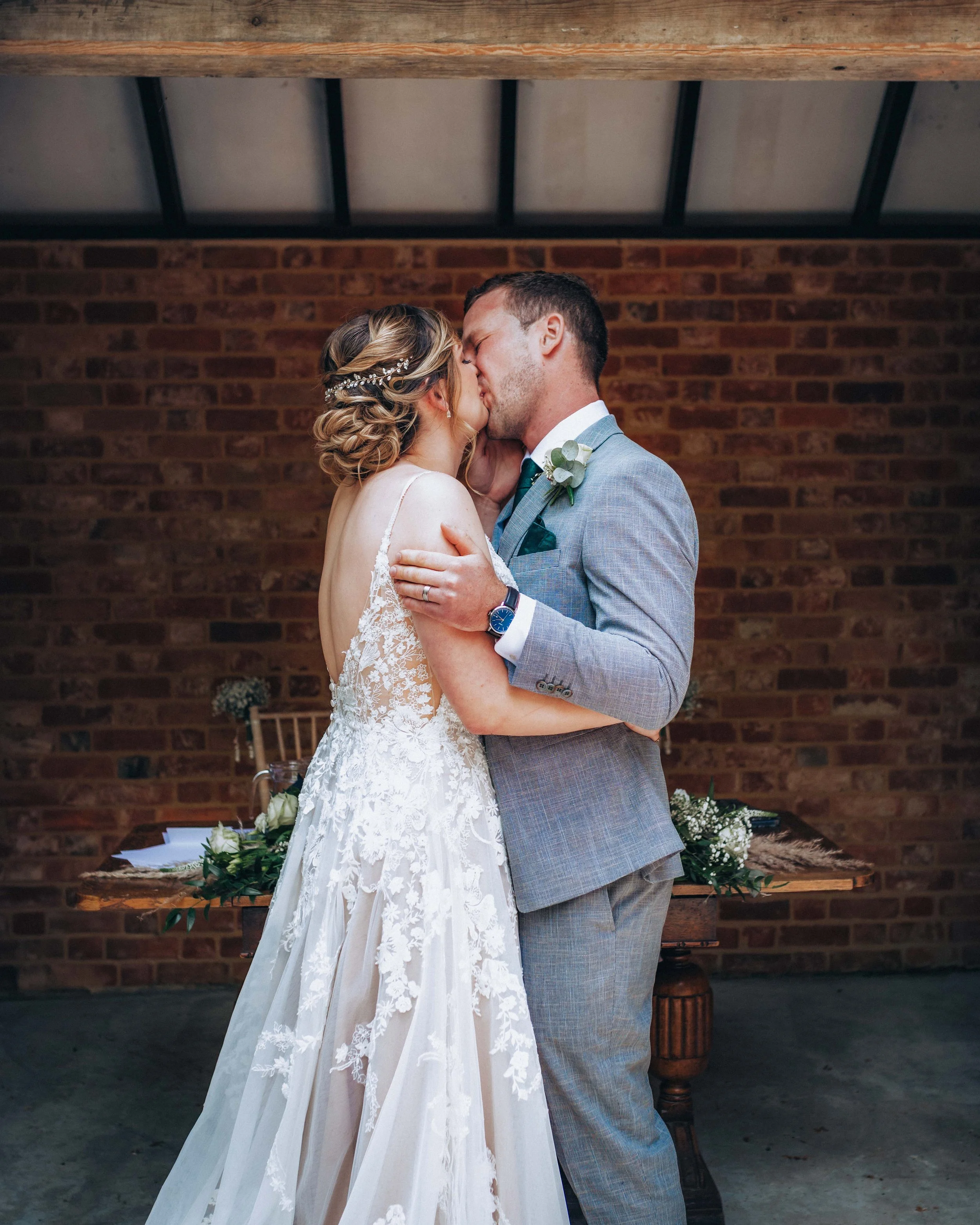 A bride and groom share a kiss during their wedding ceremony in a rustic indoor setting with a brick wall background. The bride wears a lace wedding dress and a pearl hair accessory, while the groom wears a gray suit with a white shirt and a boutonni