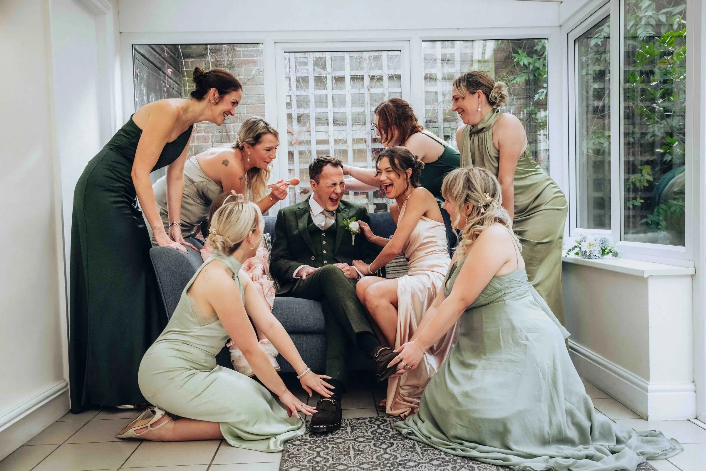 Group of women surrounding a laughing groom in a conservatory, celebrating at a wedding at East Mersea Hall Colchester.