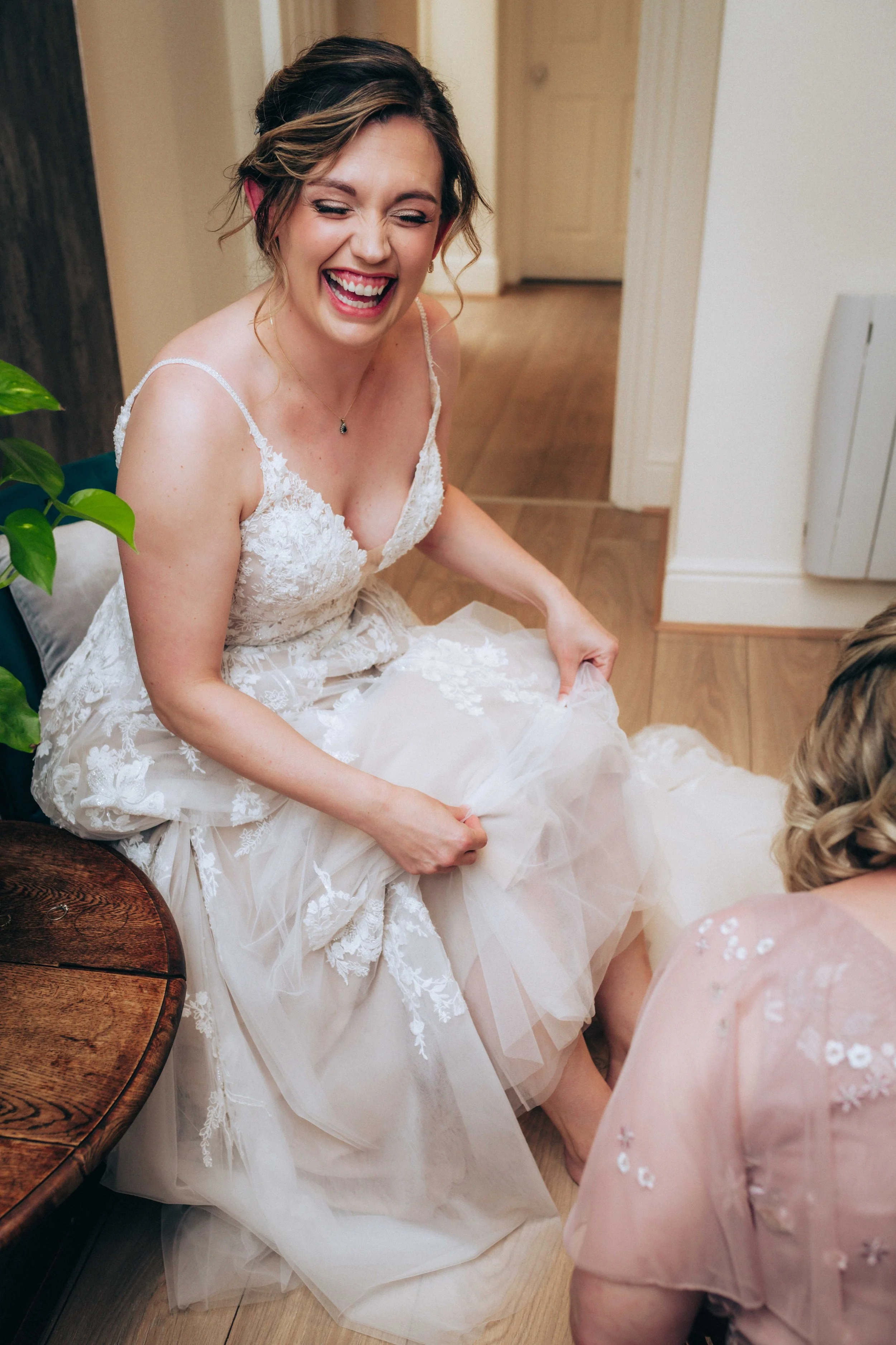 A woman in a wedding dress laughing, seated indoors near a wooden table with a plant nearby.