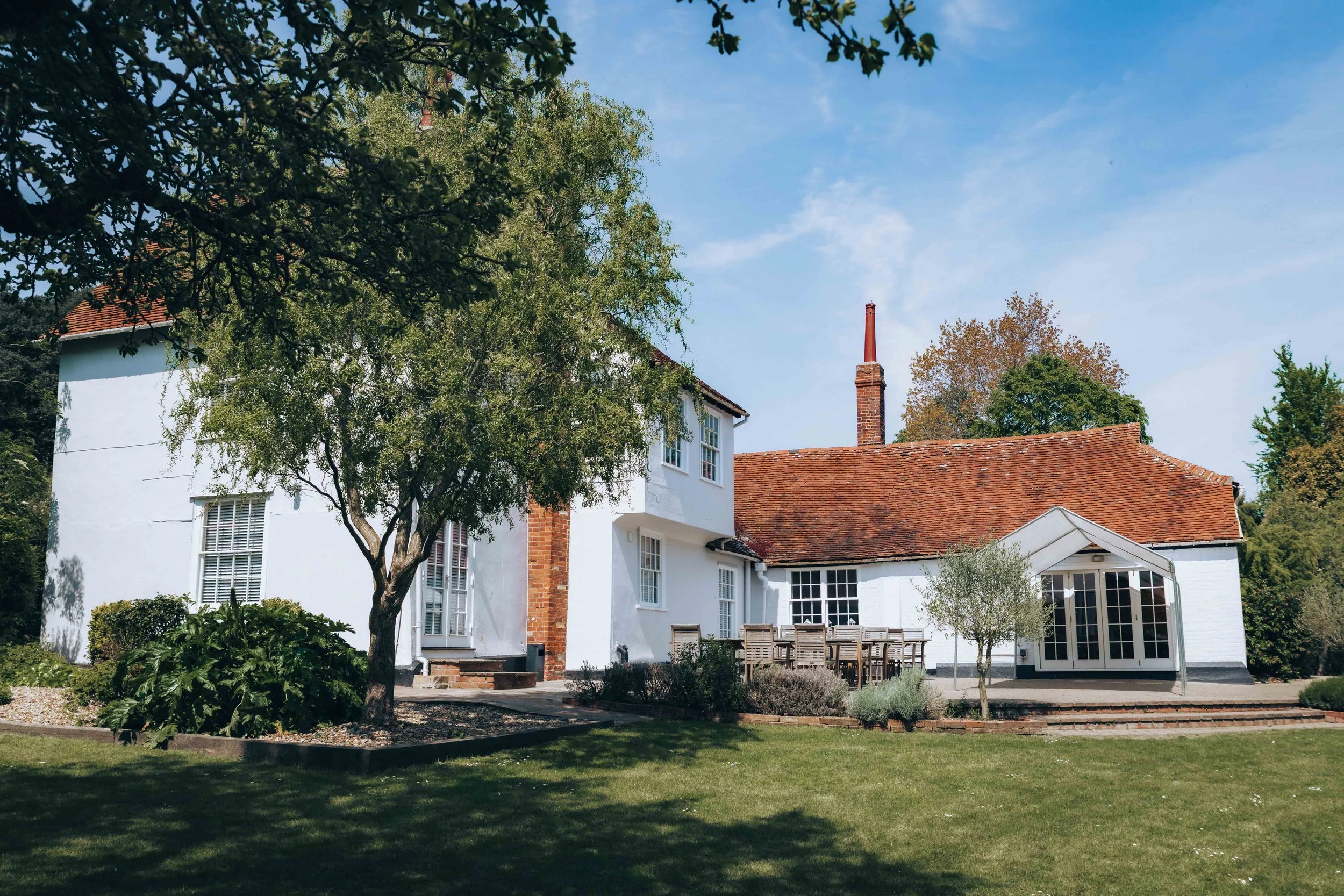 A white house with a red-tiled roof, surrounded by trees and a landscaped backyard with outdoor dining furniture.