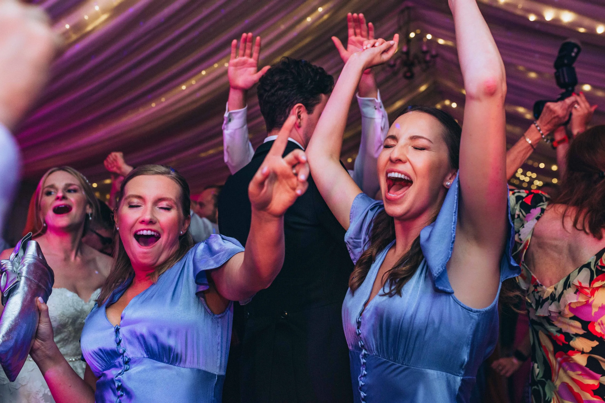 Bridesmaids with the bride in the background are dancing on the dancefloor with their arms up in the air in Essex