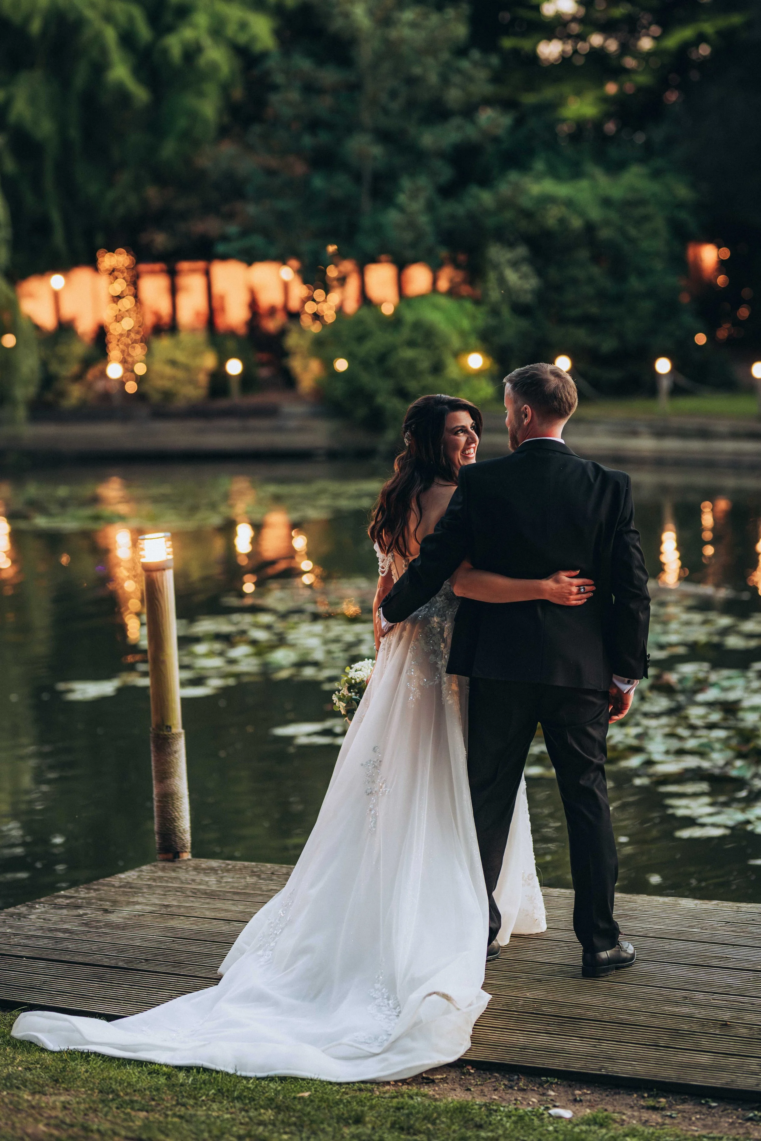 A bride and groom standing on a dock by a lake during sunset, with the bride wearing a white gown and the groom in a black suit. They are smiling at each other, with greenery and string lights in the background.