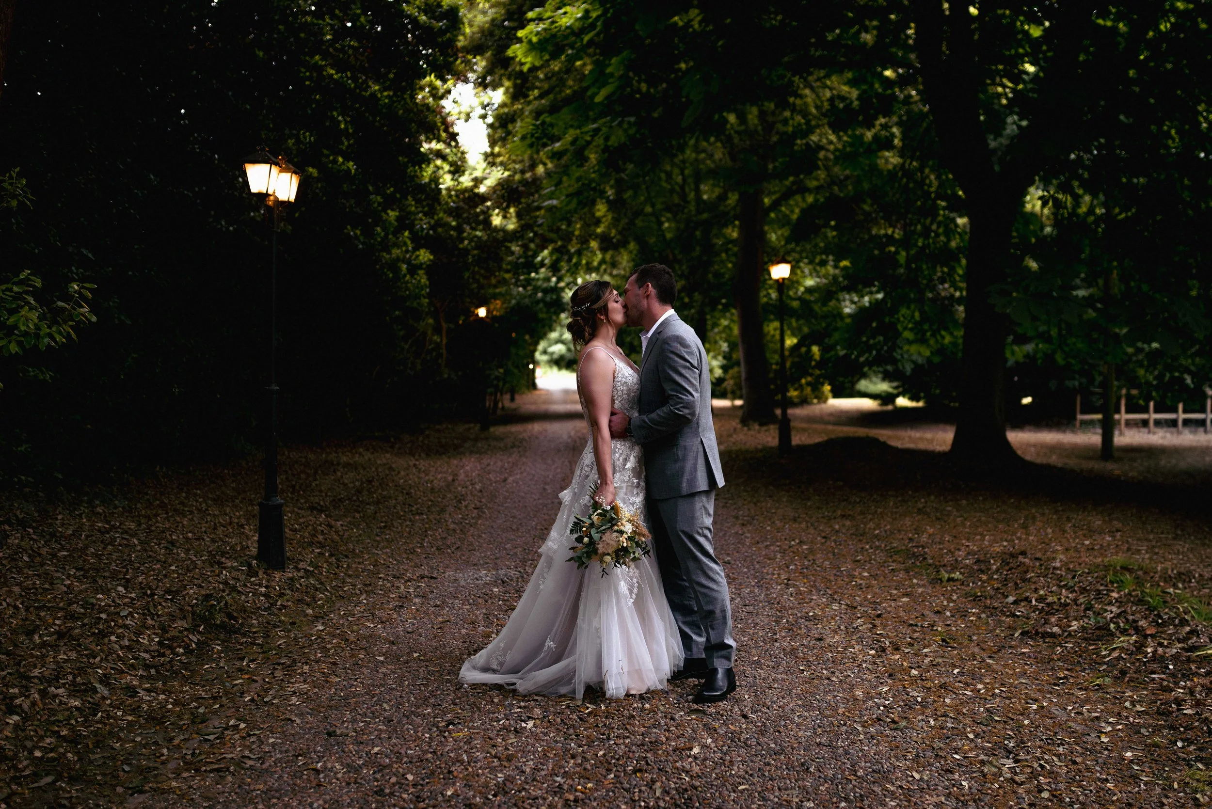 A bride and groom kiss on a tree-lined pathway at dusk, surrounded by greenery and illuminated by vintage lampposts, in a romantic outdoor setting.